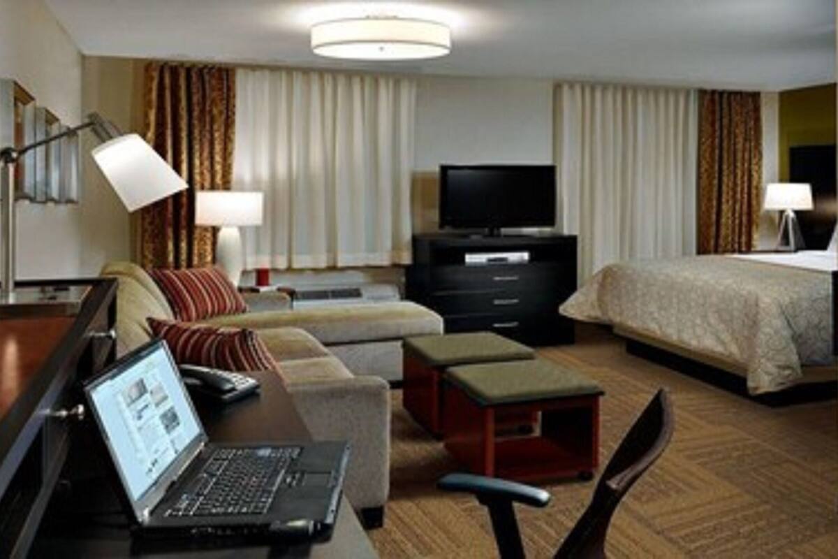 Ein Hotelzimmer mit einem Bett, einem Schreibtisch mit einem Laptop, einem Fernseher und einem Sofa.
