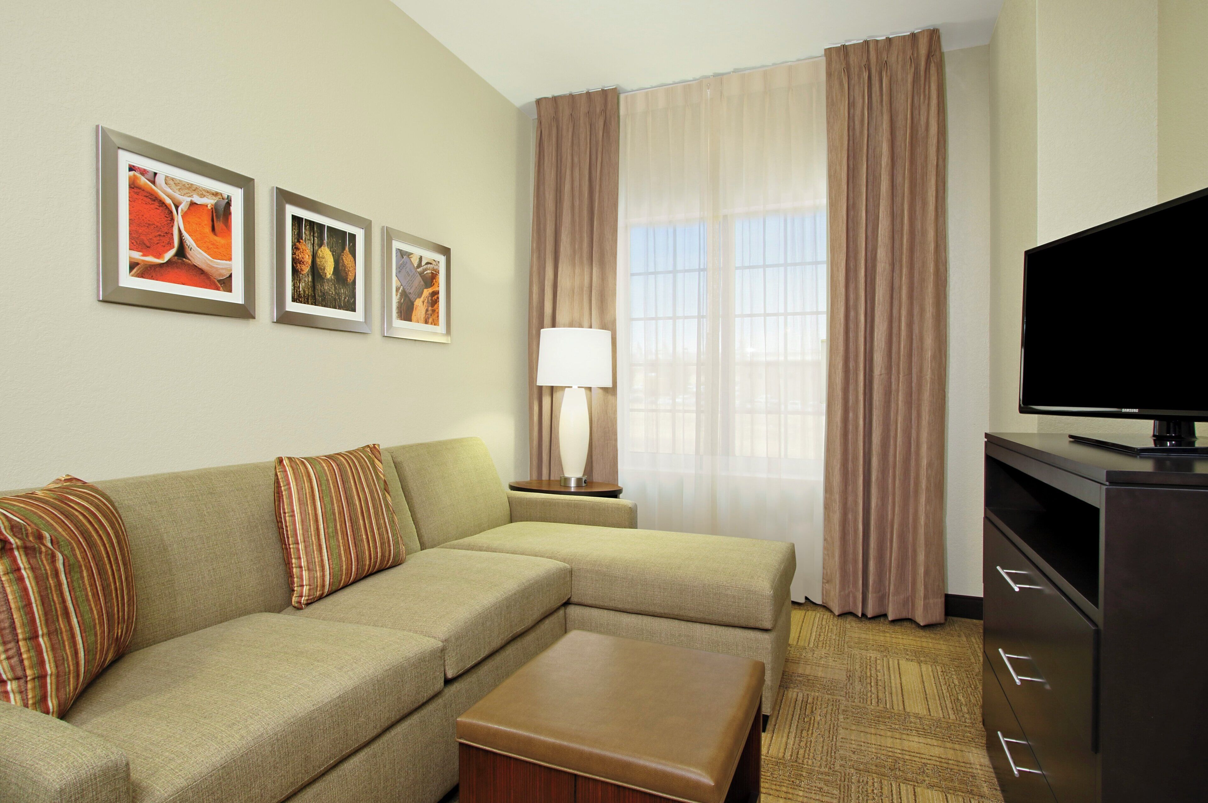 A hotel room with a sofa, a flat-screen TV, a lamp, and a window with curtains.
