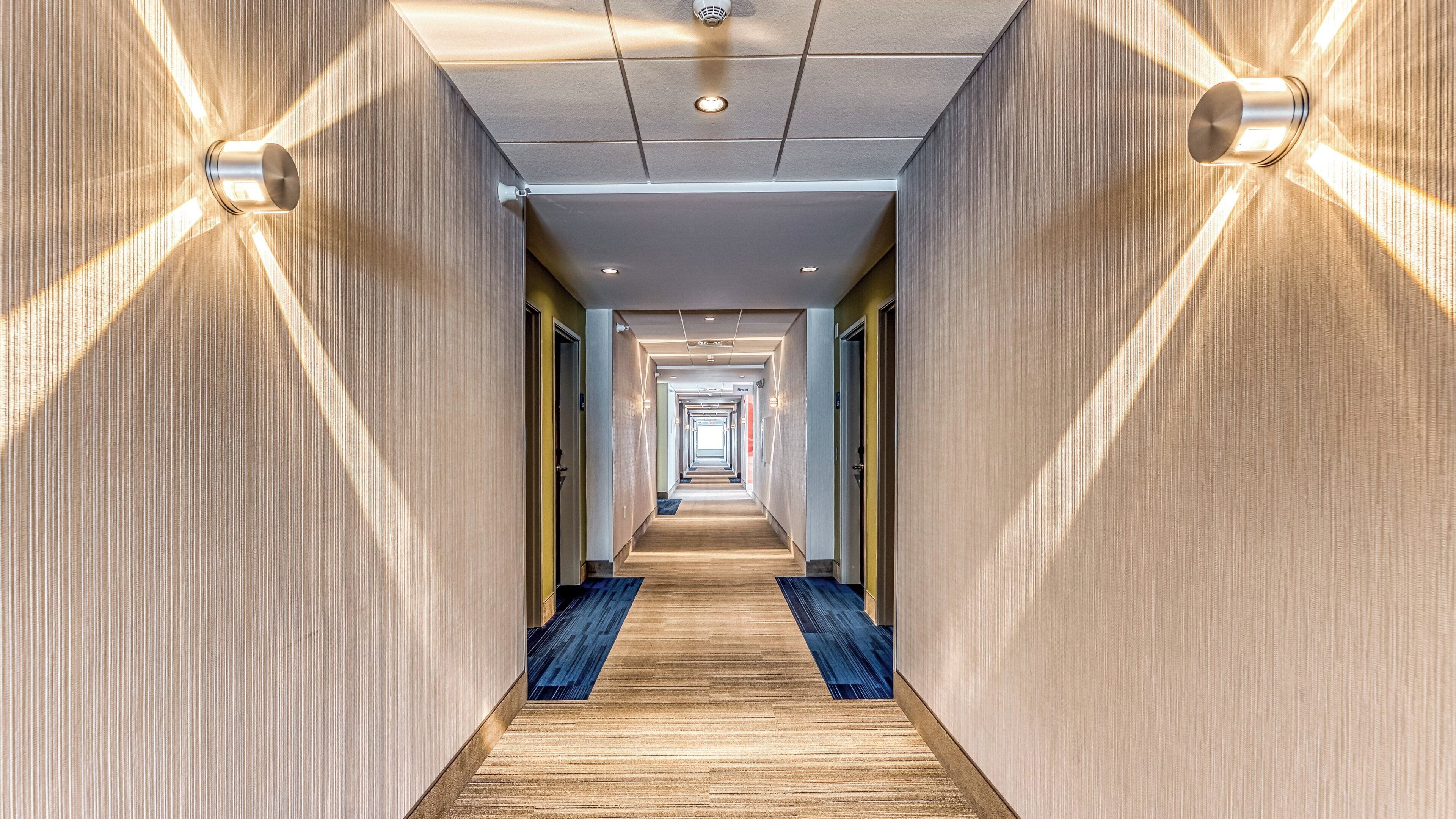 A hotel corridor with a wooden floor, light-colored walls, and a ceiling with recessed lighting.