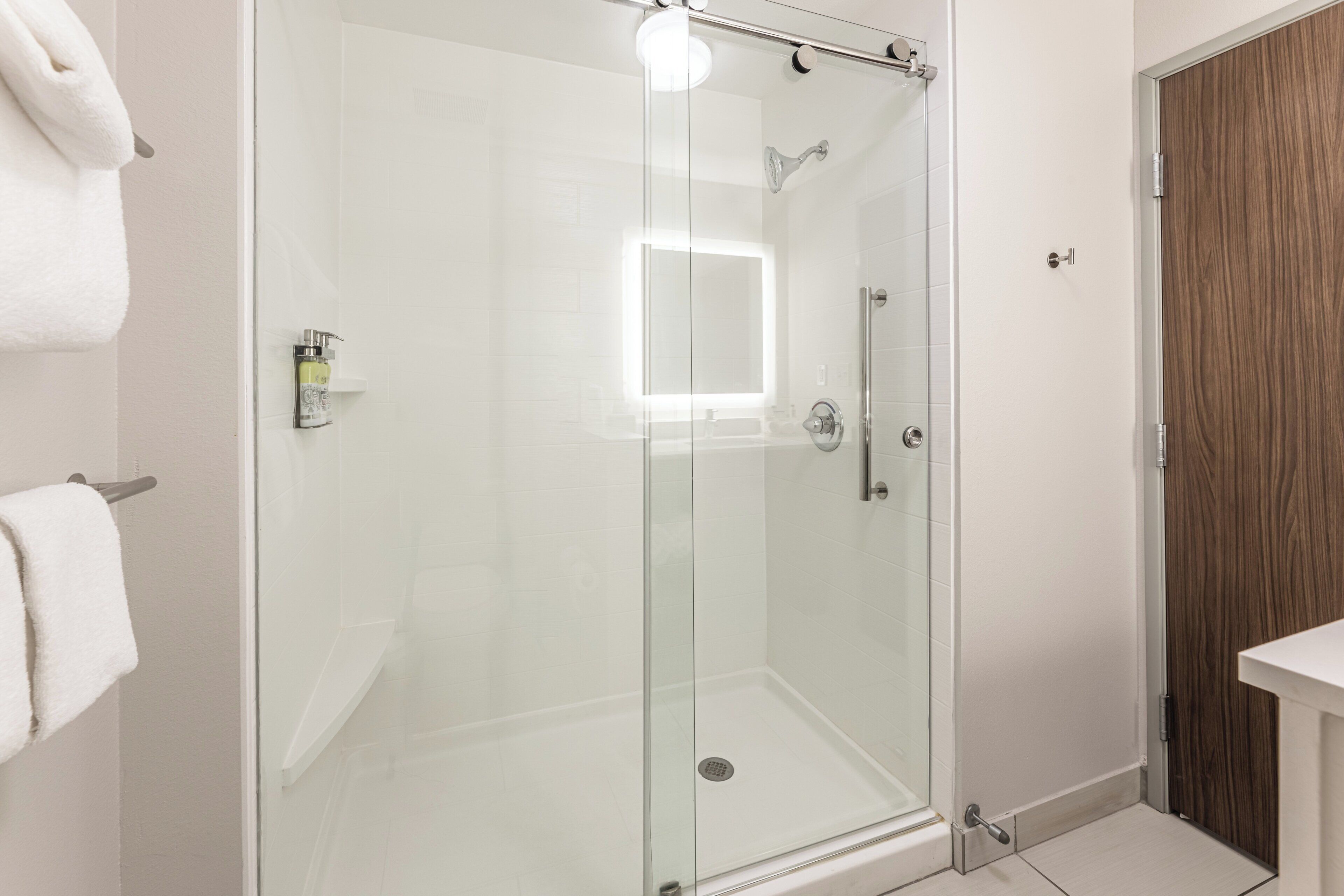 A glass-enclosed shower stall with a white tiled wall, a wooden door, and a white towel on a towel rack.