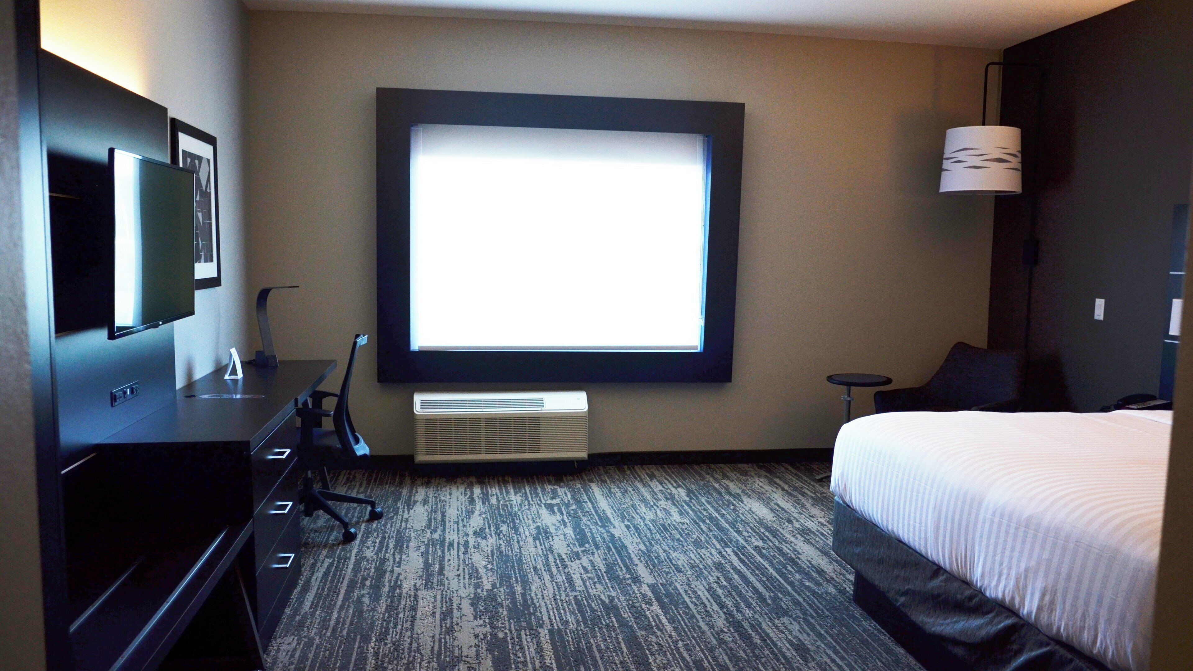 Habitación de hotel con un ventanal grande, cama, escritorio con silla, televisor y lámpara.