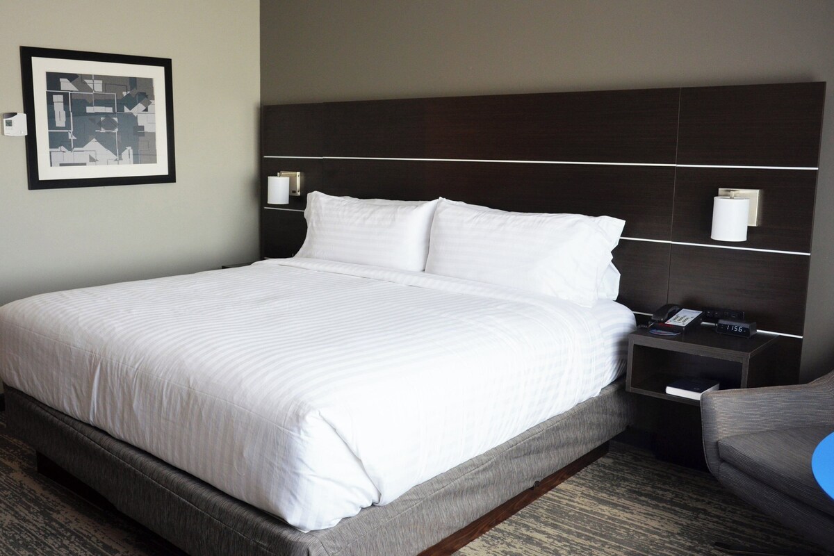 Une chambre d’hôtel avec un grand lit, une tête de lit, des tables de chevet, une chaise et un tableau encadré au mur.