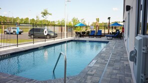 Una piscina rectangular con una escalera metálica, rodeada por una valla de metal negro. Hay sombrillas azules y sillas de descanso junto a la piscina.