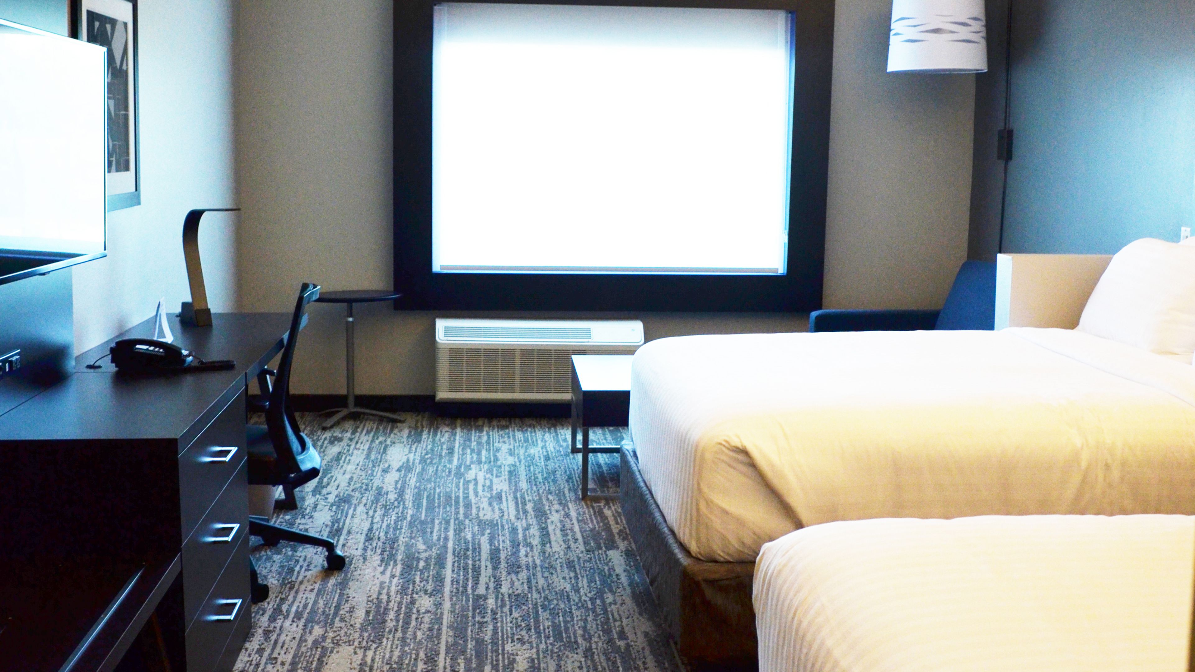 A hotel room with a bed, a desk with a phone and a lamp, a TV, and a window.