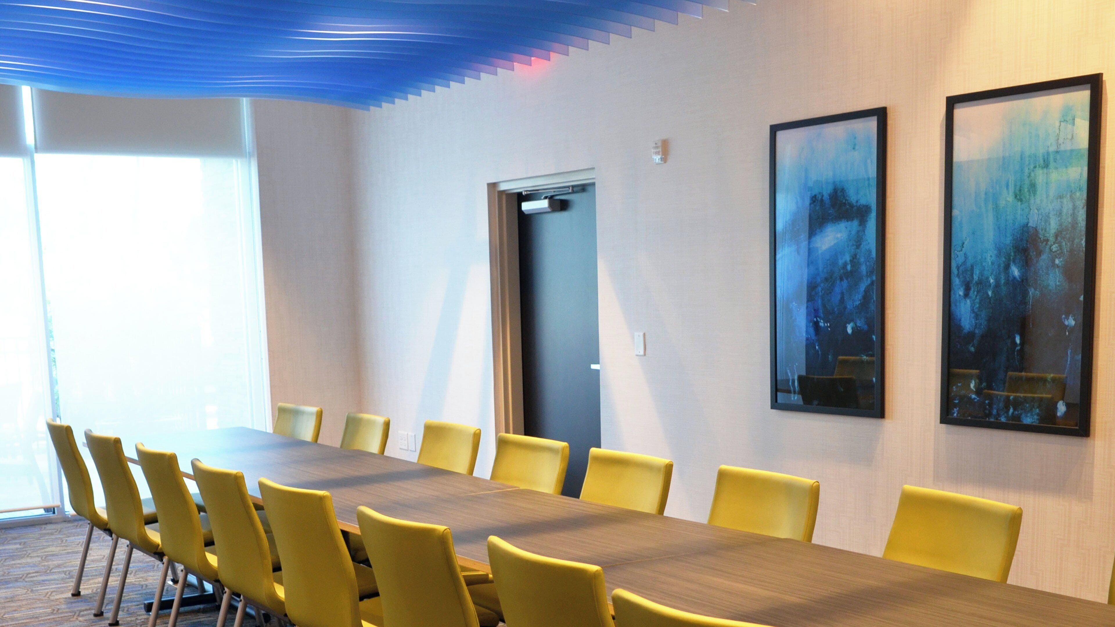 A conference room with a long table and yellow chairs, a door, and two framed artworks on the wall.