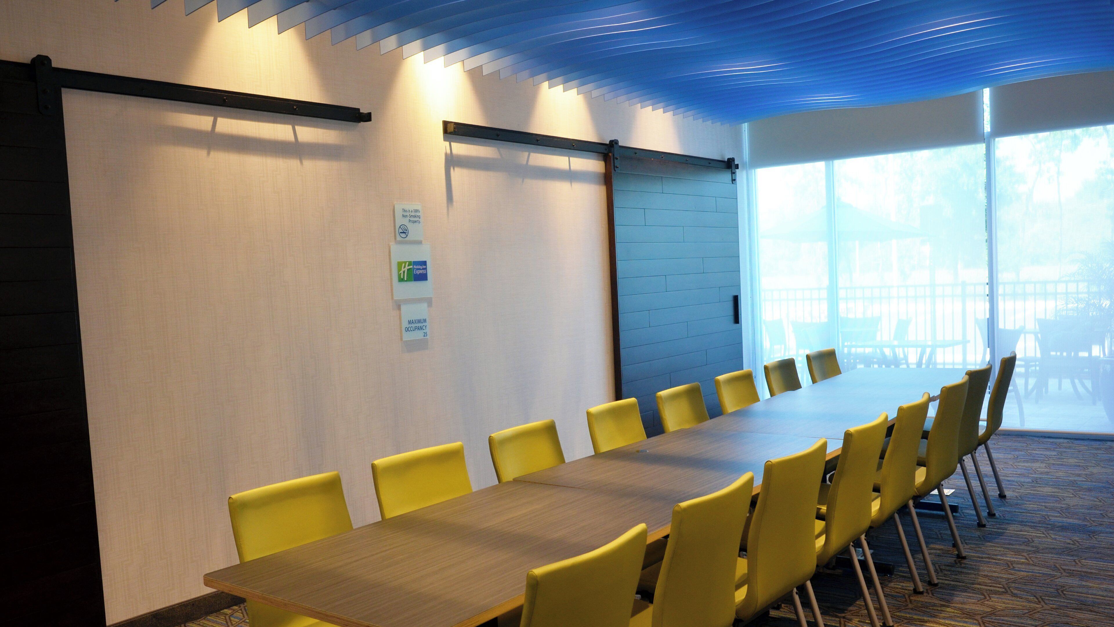A conference room with a long table and yellow chairs, a whiteboard, and a window with a view of outdoor furniture.