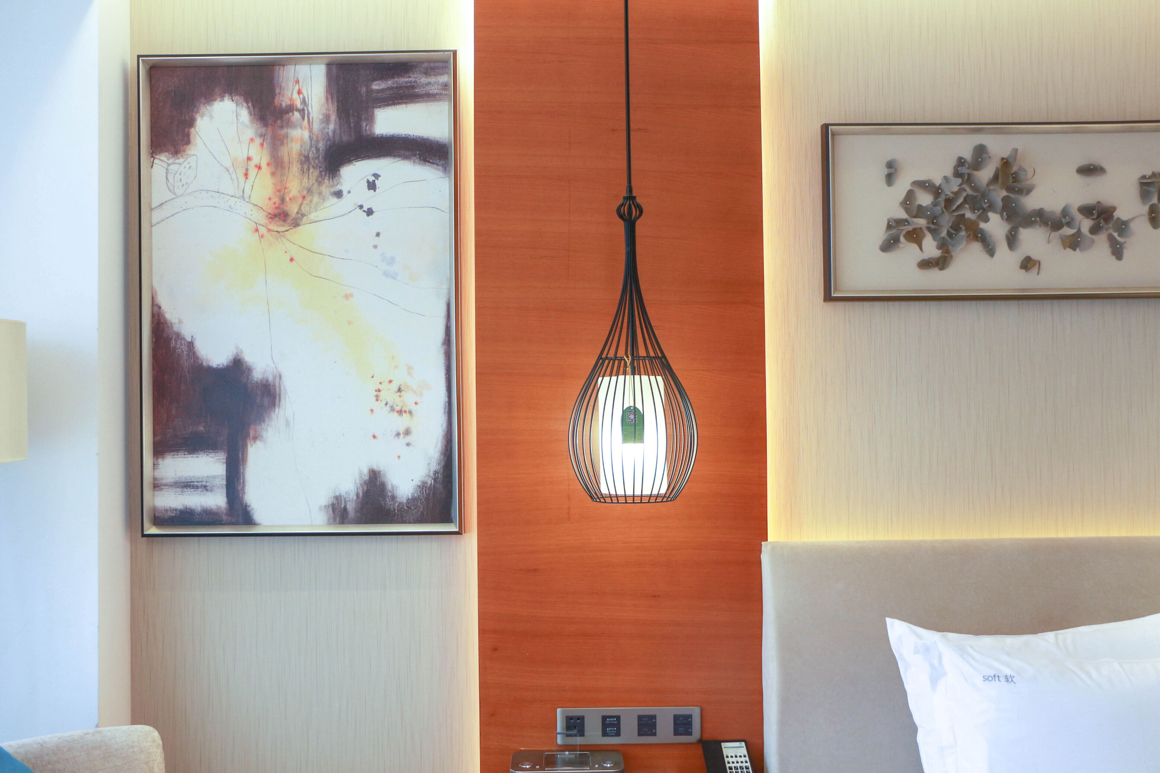 A hotel room with a large abstract painting, a hanging pendant light, a control panel, and a bed with a white pillow.