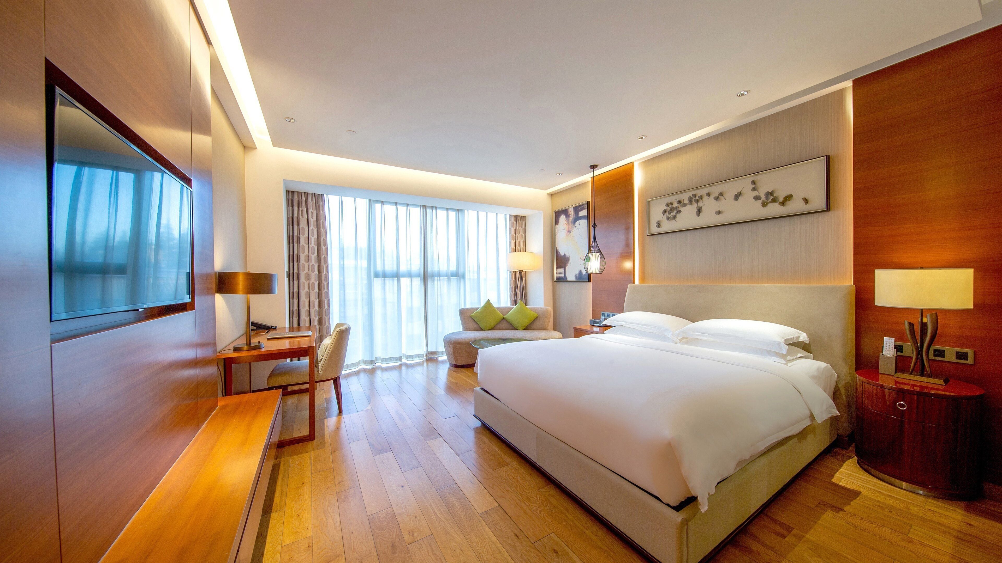 A modern hotel room with a large bed, a desk, a chair, a TV, and a large window with curtains.