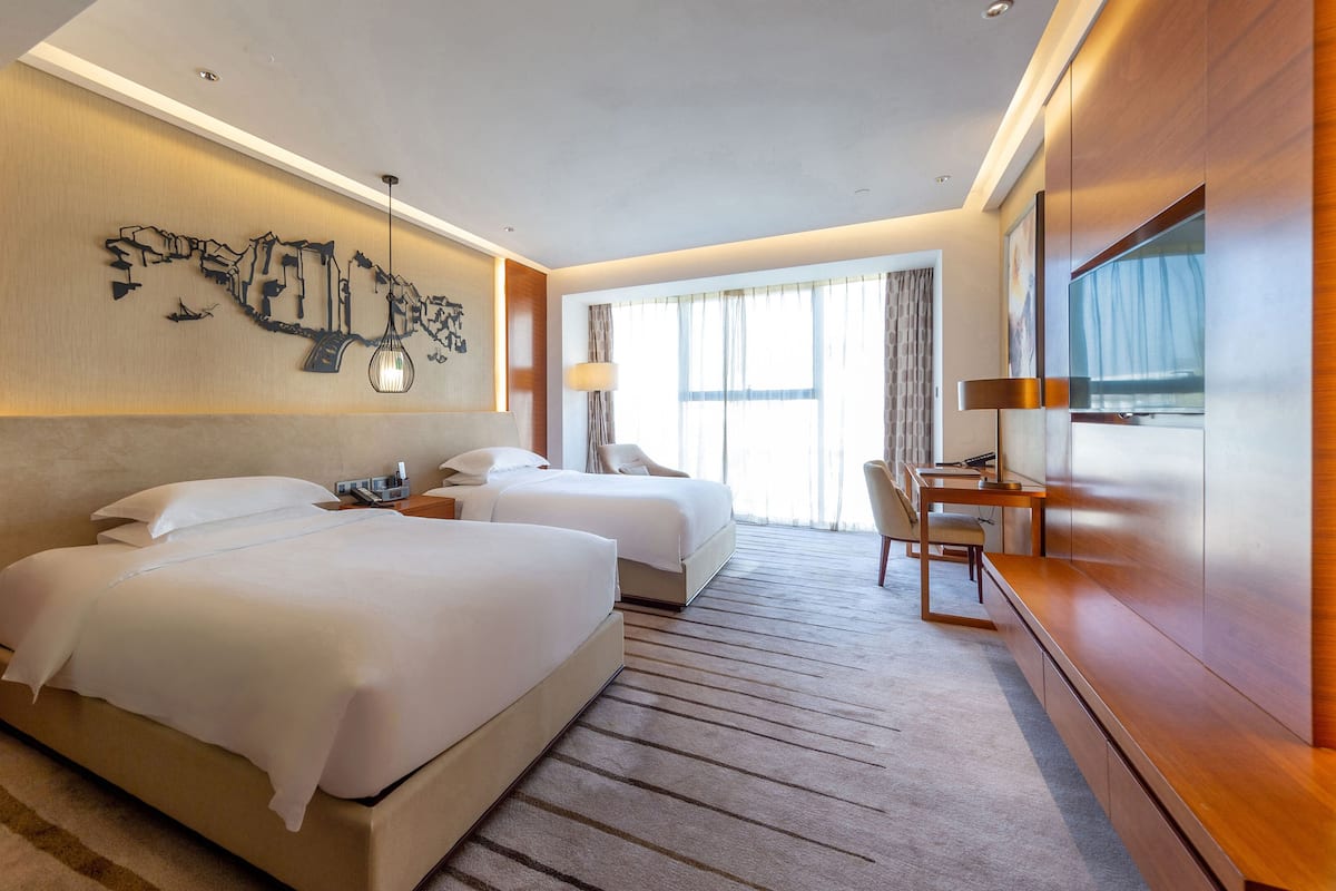 A hotel room with two beds, a desk, a chair, a large window, and a wall-mounted artwork.