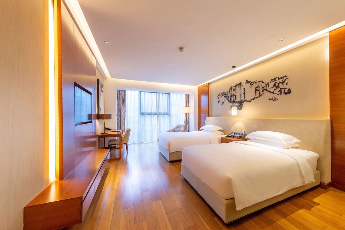 A modern hotel room with two beds, a desk, and a large window with curtains.