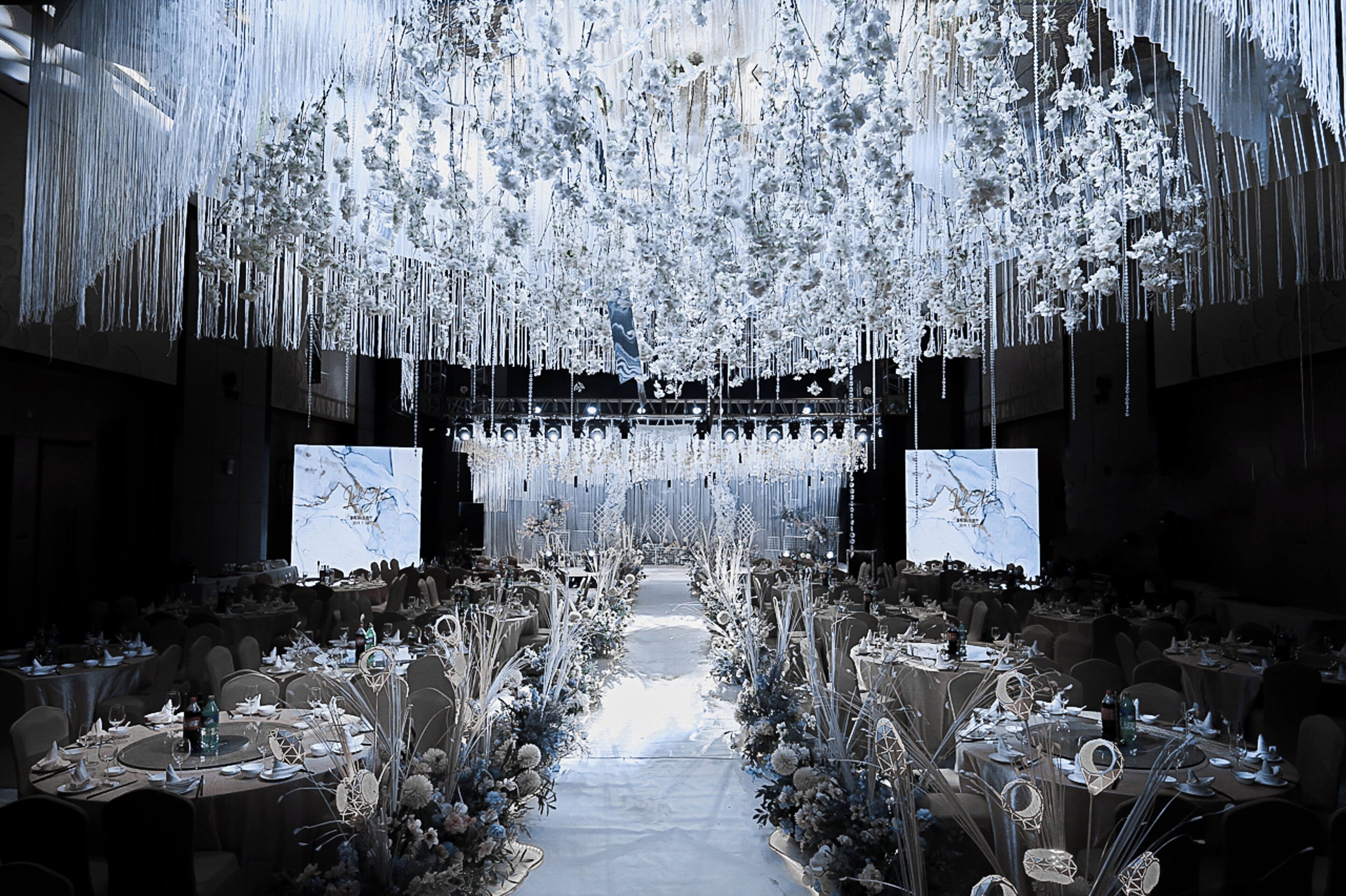 A banquet hall with tables set for a formal event, a central aisle, and hanging floral decorations.