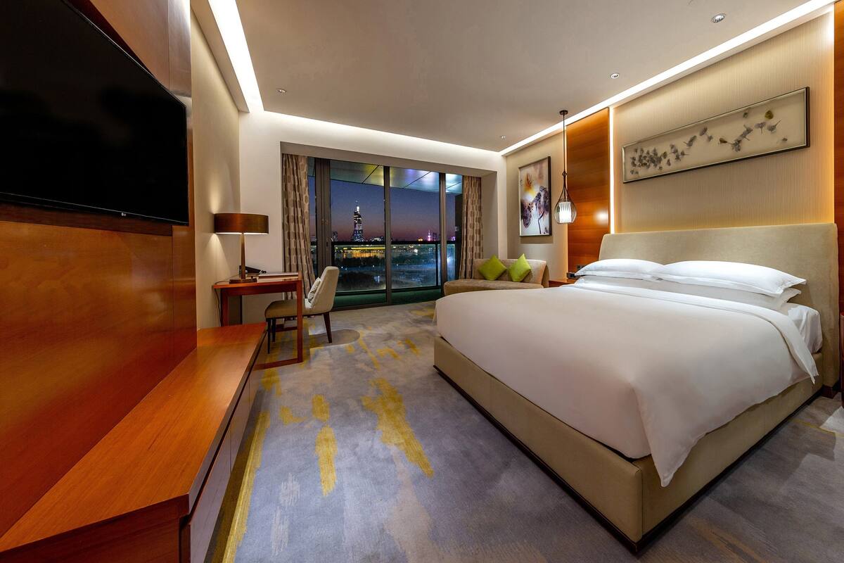 A hotel room with a large bed, a desk, a chair, a TV, and a view of the cityscape.