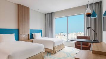Habitación de hotel con dos camas, un escritorio, una silla, un ventanal grande con vistas a la ciudad y una lámpara moderna.