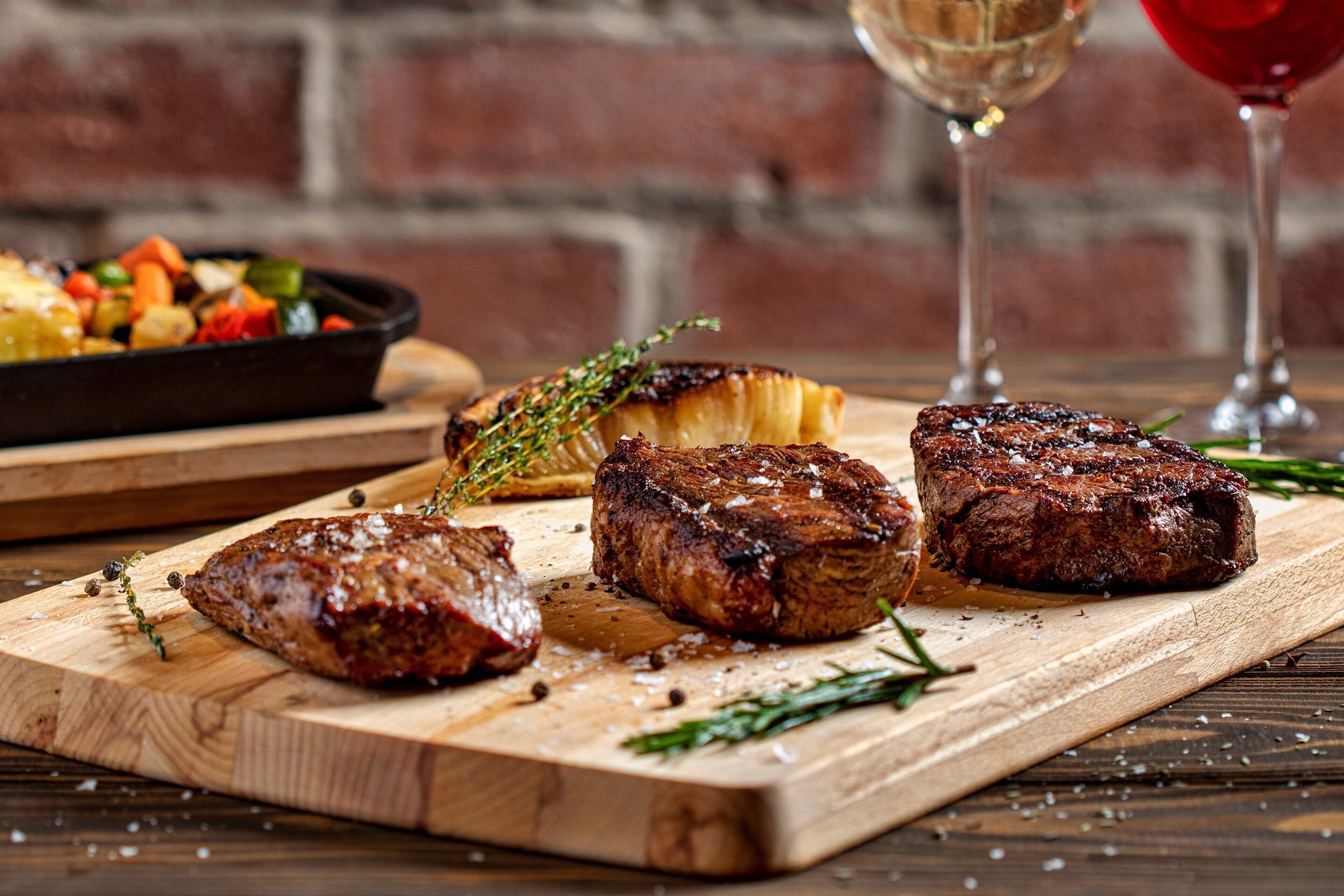 Una tabla de madera con bistec y verduras, una copa de vino tinto, y un fondo de pared de ladrillo.