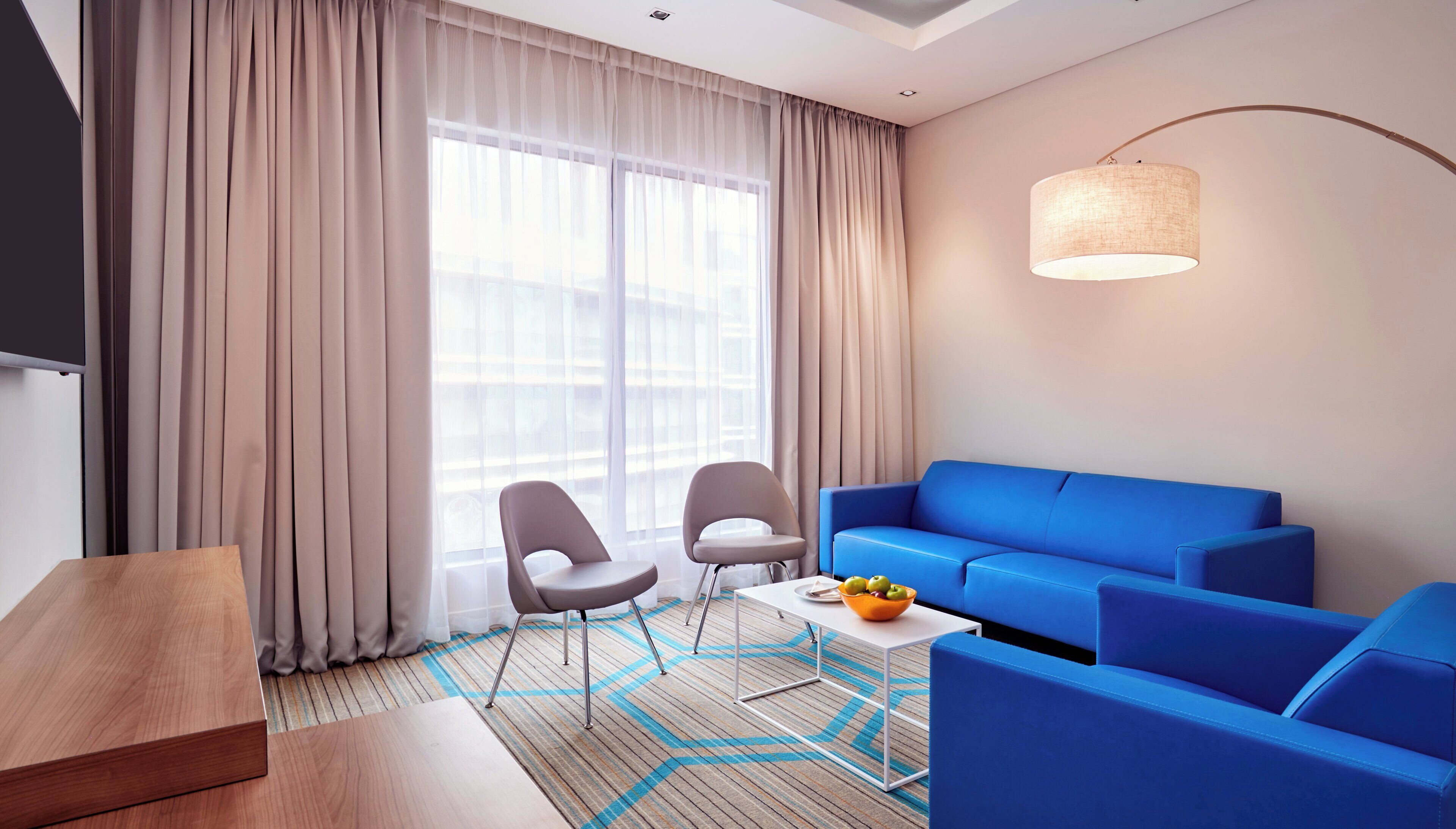 Habitació d'hotel moderna amb un sofà blau, dues cadires, una taula petita amb fruita i una televisió.