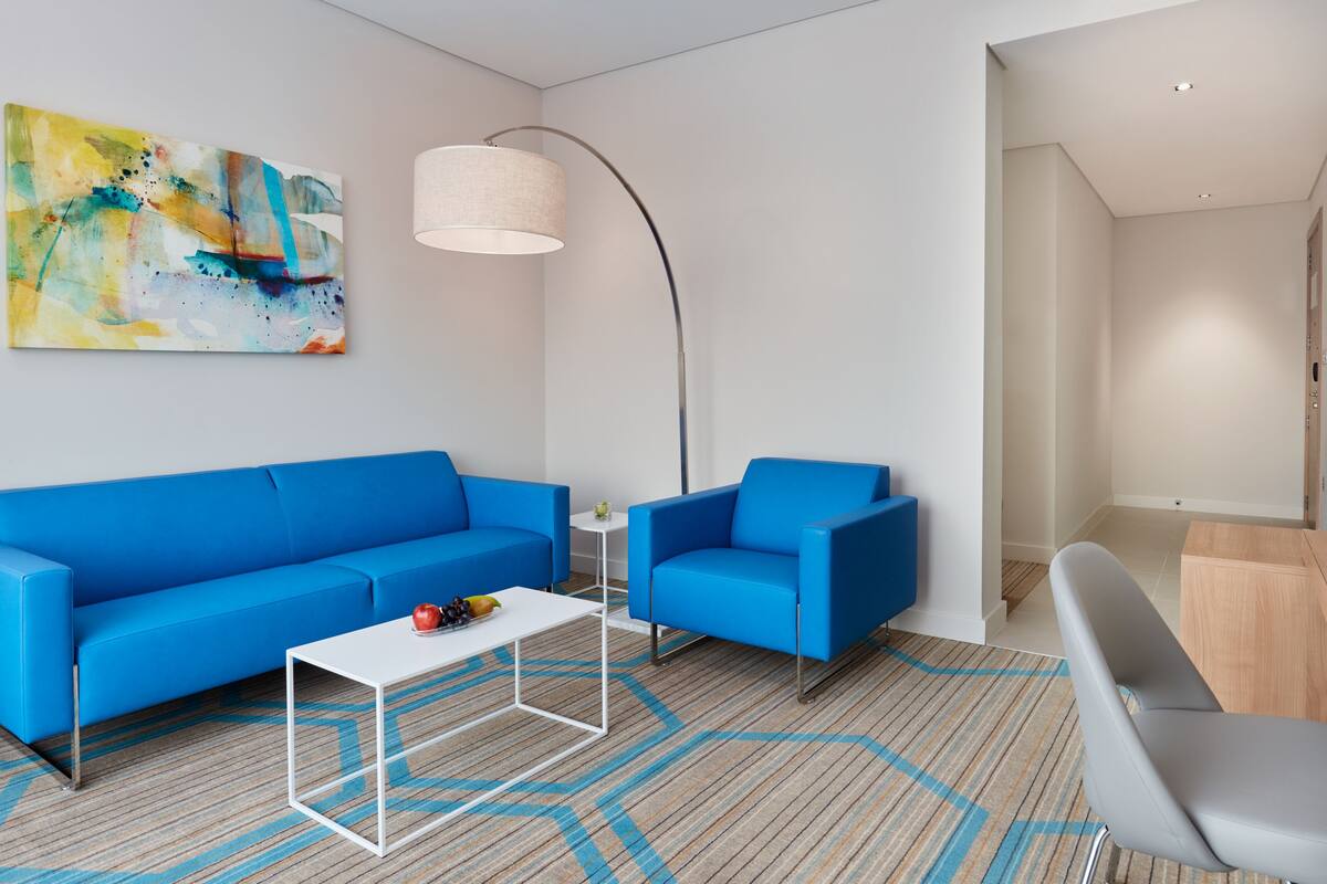 A modern living room with a blue sofa, a white coffee table, and a grey armchair. There's a floor lamp and a painting on the wall.