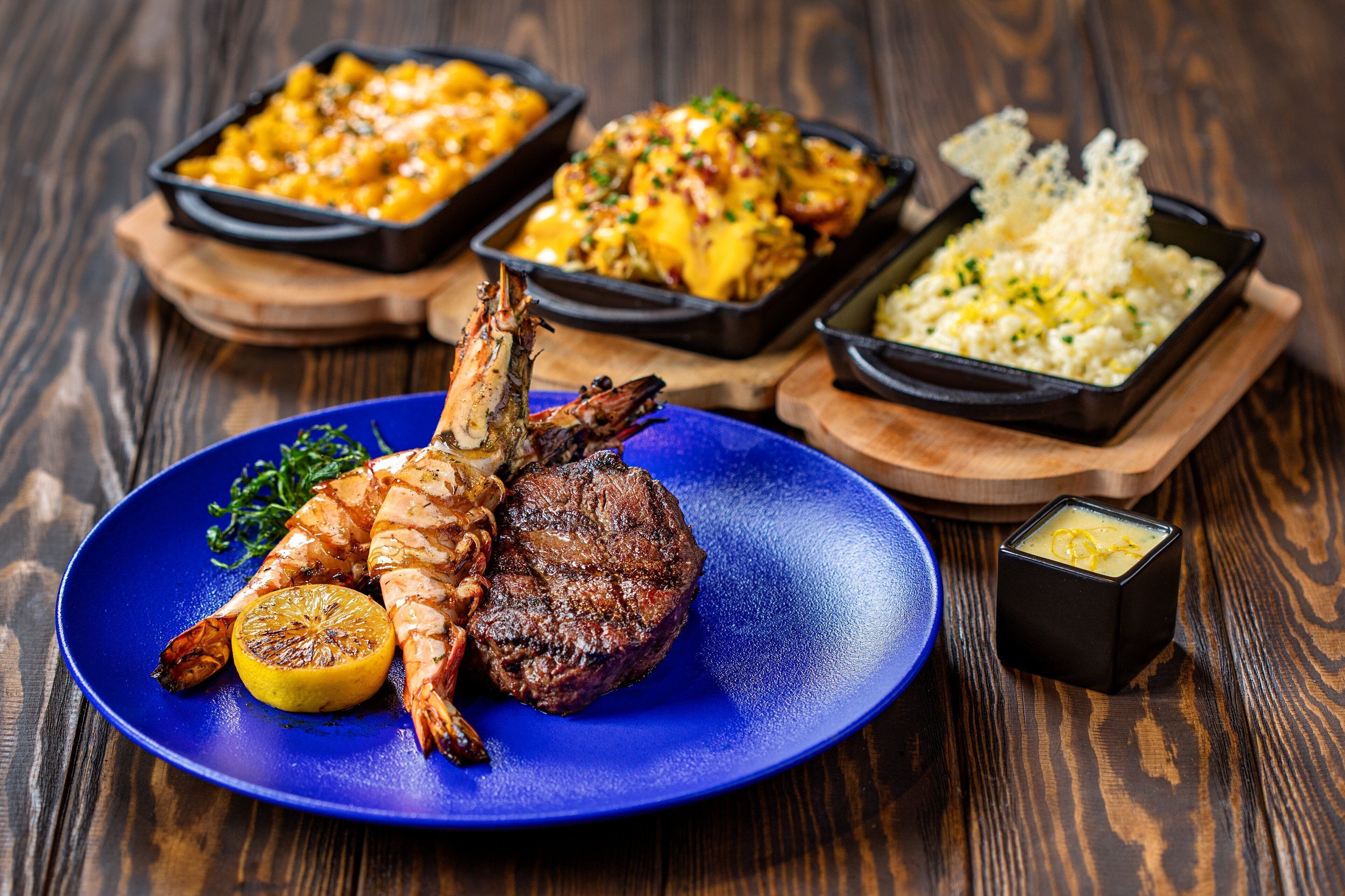 A plate with grilled fish, steak, and lemon on a blue plate, accompanied by three side dishes in black containers.