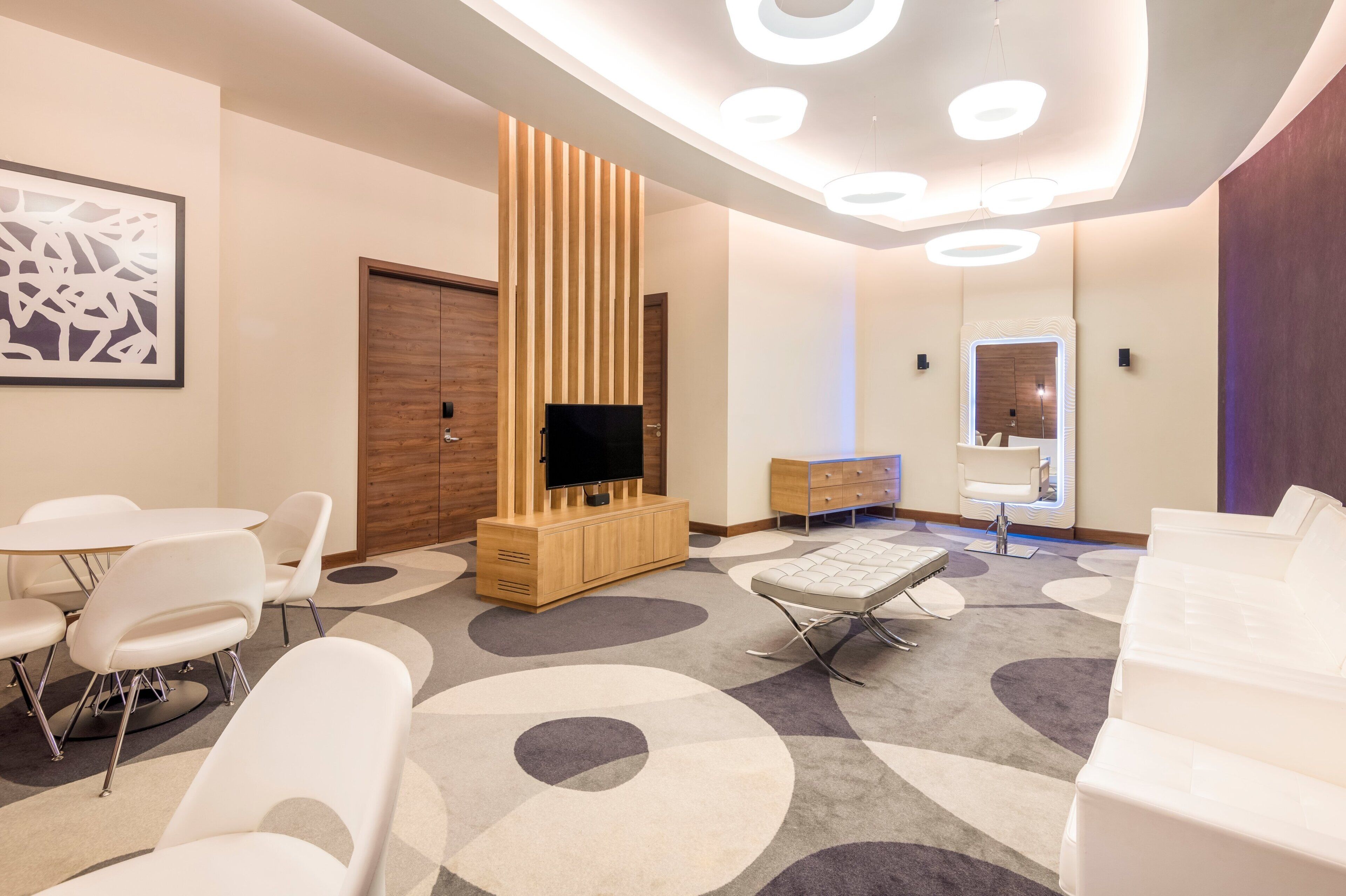 A modern hotel room with a flat-screen TV, a wooden cabinet, a sofa, a dining table with chairs, and a patterned carpet.