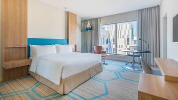 A hotel room with a large bed, a desk, a chair, and a view of the cityscape.