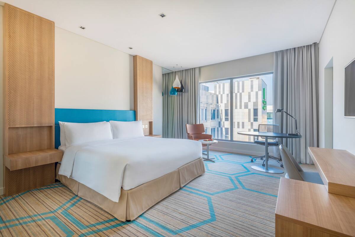 A hotel room with a large bed, a desk with a chair, a small table, and a large window with a view of buildings.