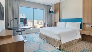 A hotel room with a large bed, a desk, a chair, a lamp, and a view of the city through large windows.