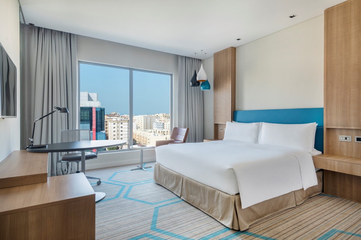 Une chambre d’hôtel avec un grand lit, un bureau, une chaise, une lampe et une vue sur la ville grâce à de grandes fenêtres.