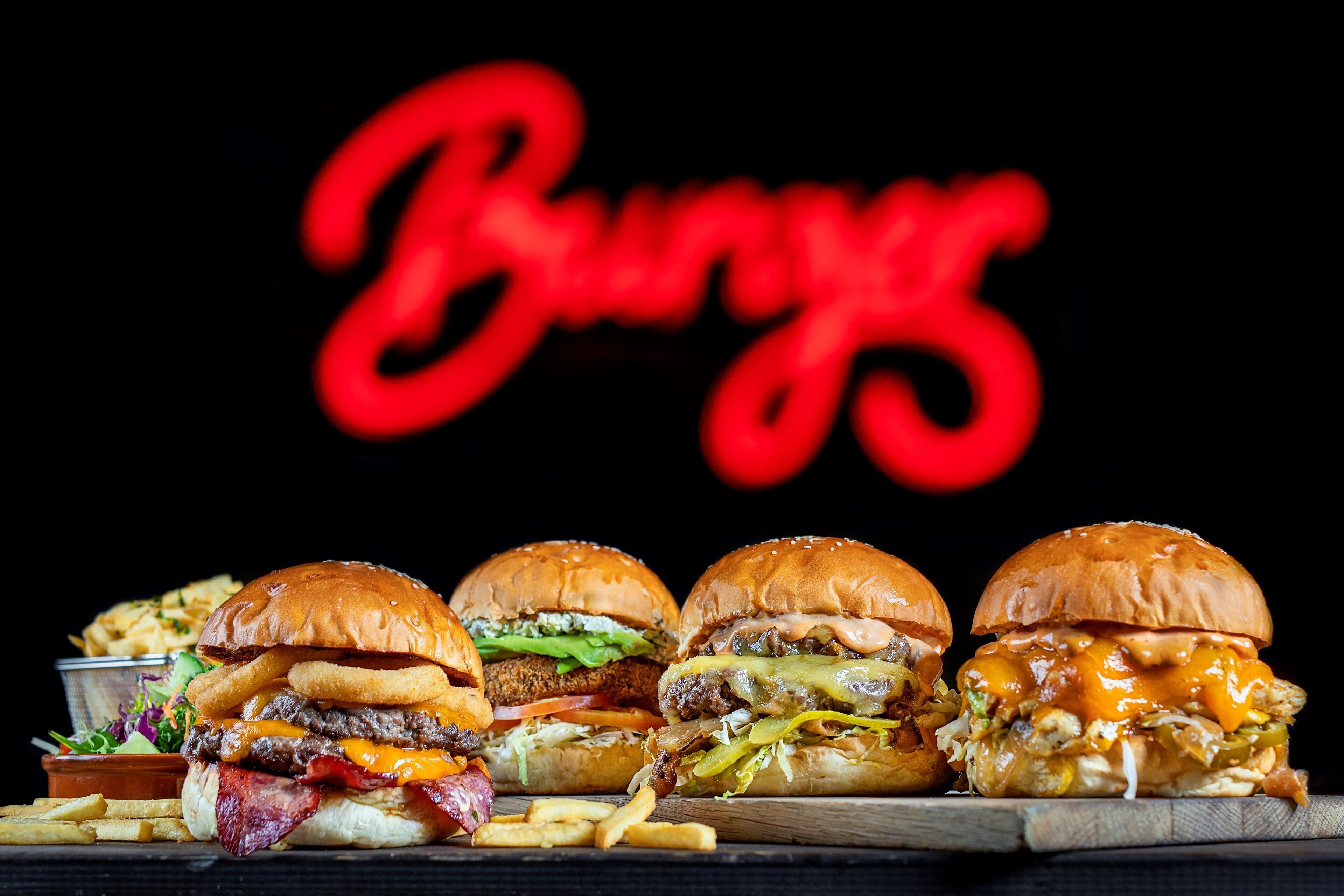 Four burgers with fries on a wooden board, and a neon sign in the background.