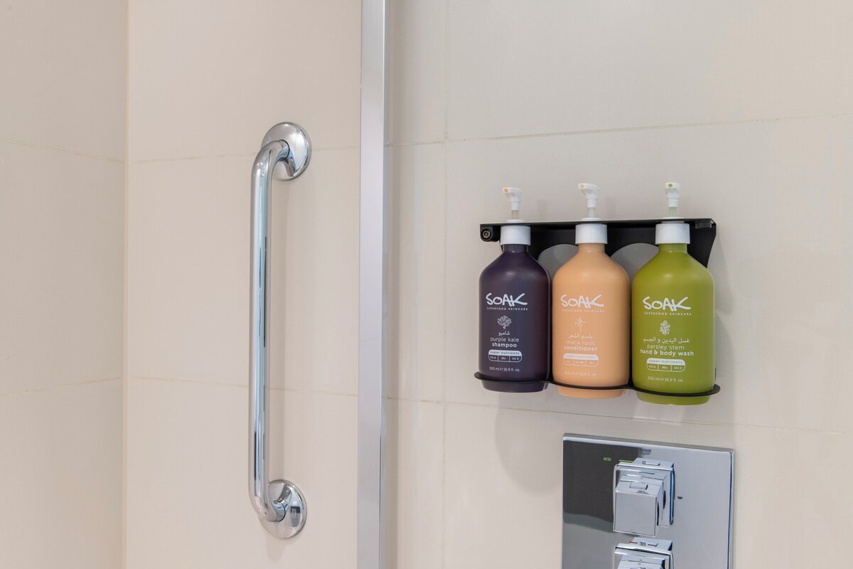 A bathroom with a handrail, three bottles of Soak body wash, and a control panel.