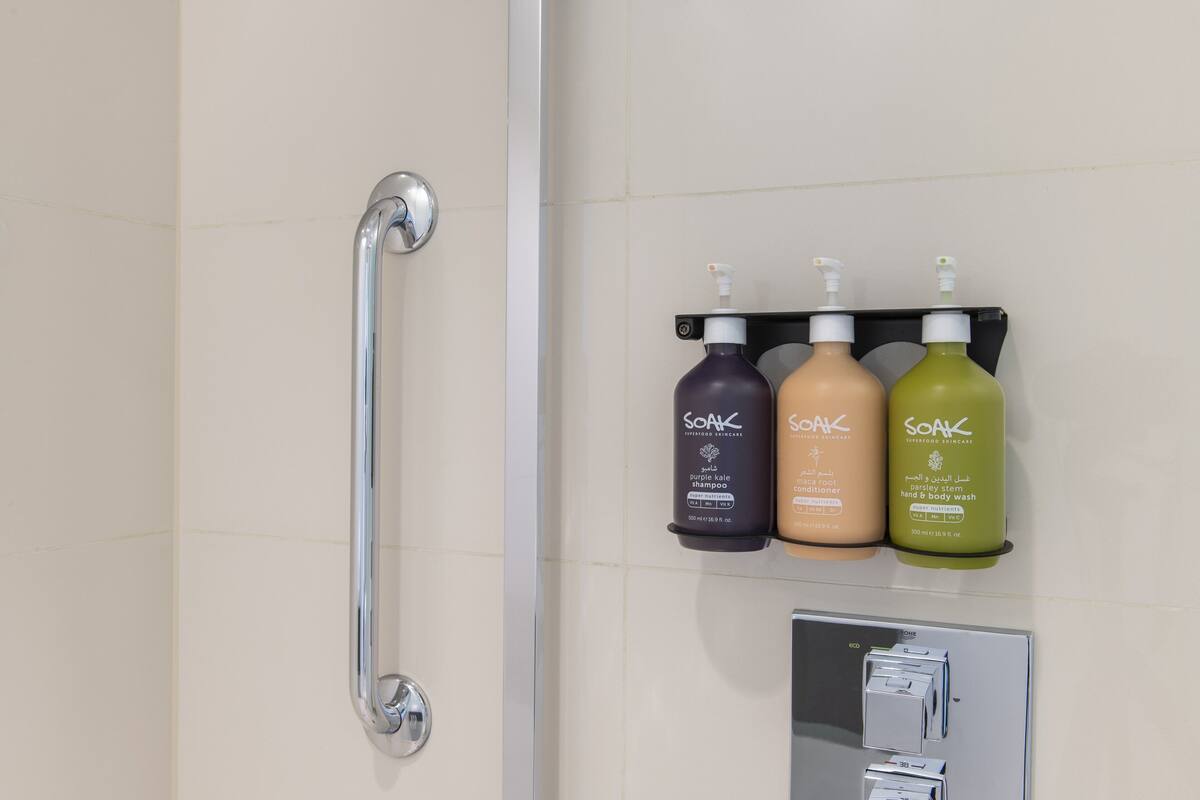 A bathroom with a handrail, three bottles of Soak body wash, and a control panel.