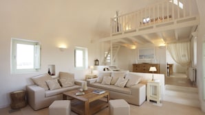 Amplia sala de estar con techo alto, una escalera que lleva a un nivel superior y vistas al exterior a través de grandes ventanales.
