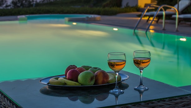 Mesa junto a la piscina con una bandeja de frutas y dos copas de vino, rodeada por una piscina y sillas de descanso.
