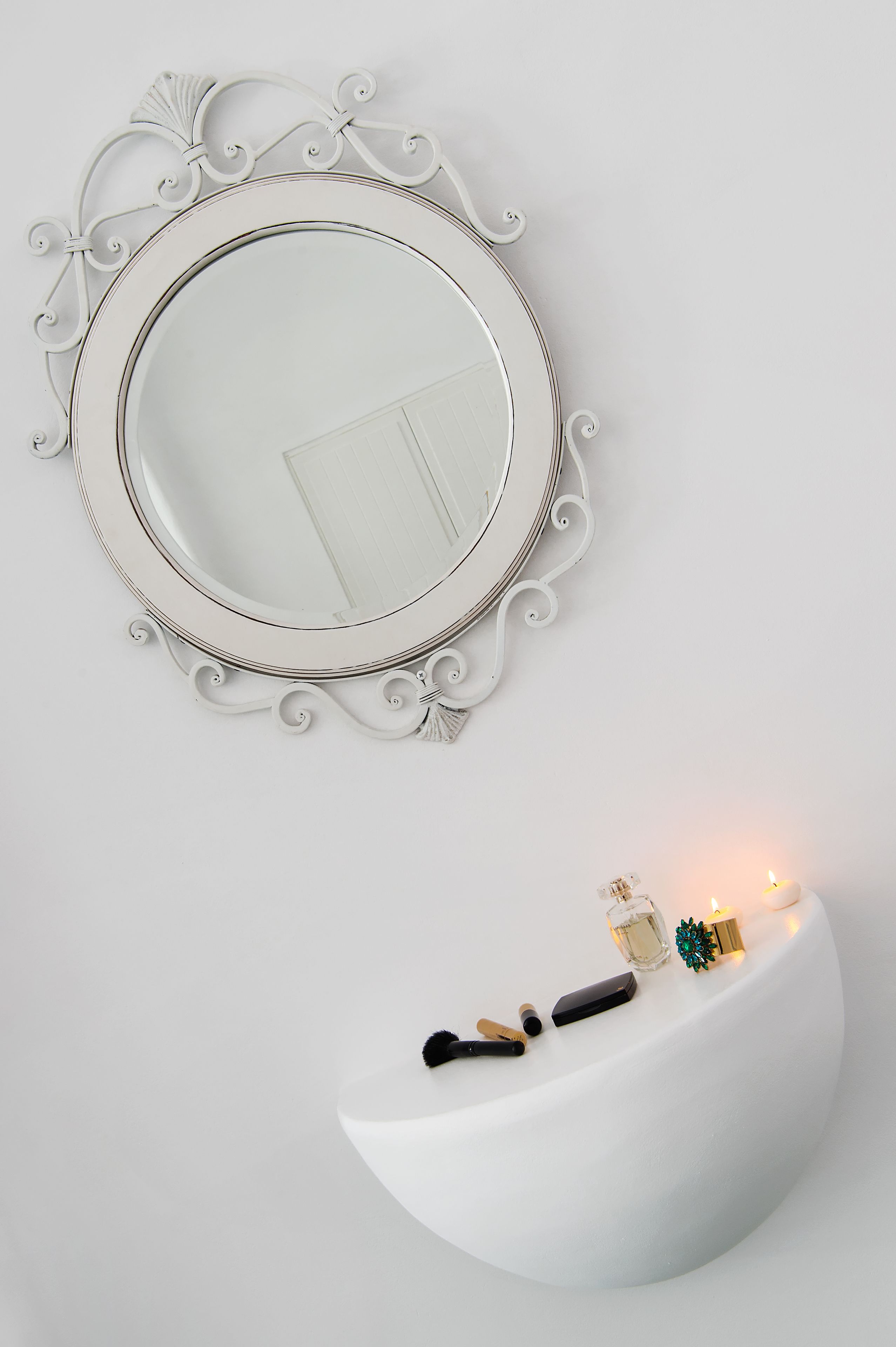 A white wall with a decorative mirror and a white shelf with a candle, a small bottle, and a decorative item.