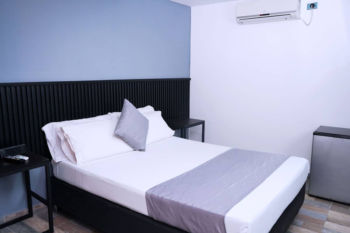 Una cama con cabecera negra, ropa de cama blanca, almohada gris, mesita de noche con un control remoto y un armario pequeño gris.