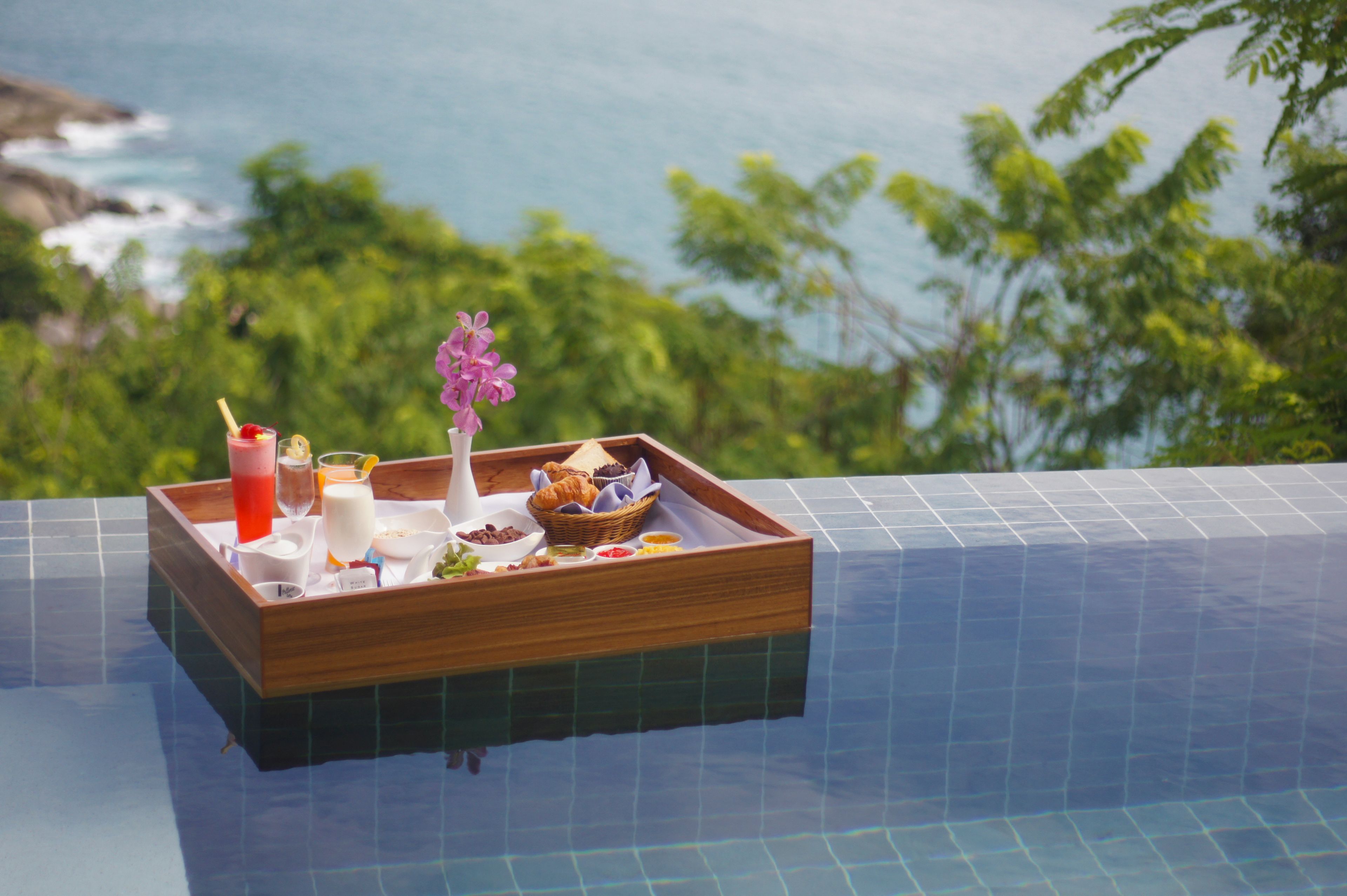 A tray with food and drinks on a poolside table with a view of the ocean and greenery.