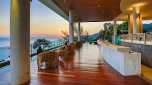 A wooden deck with wicker furniture, a marble bar, and a view of the ocean at sunset.