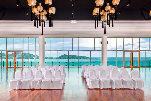 A spacious hall with rows of white chairs, large windows with a view of the sea and hills, and hanging chandeliers.