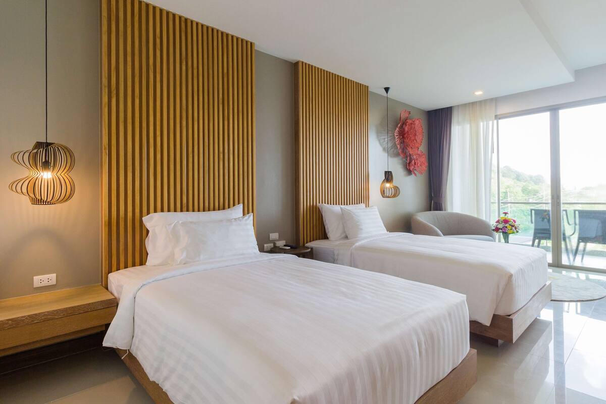 A hotel room with two beds, a wooden headboard, a pendant light, and a view of the outdoors.
