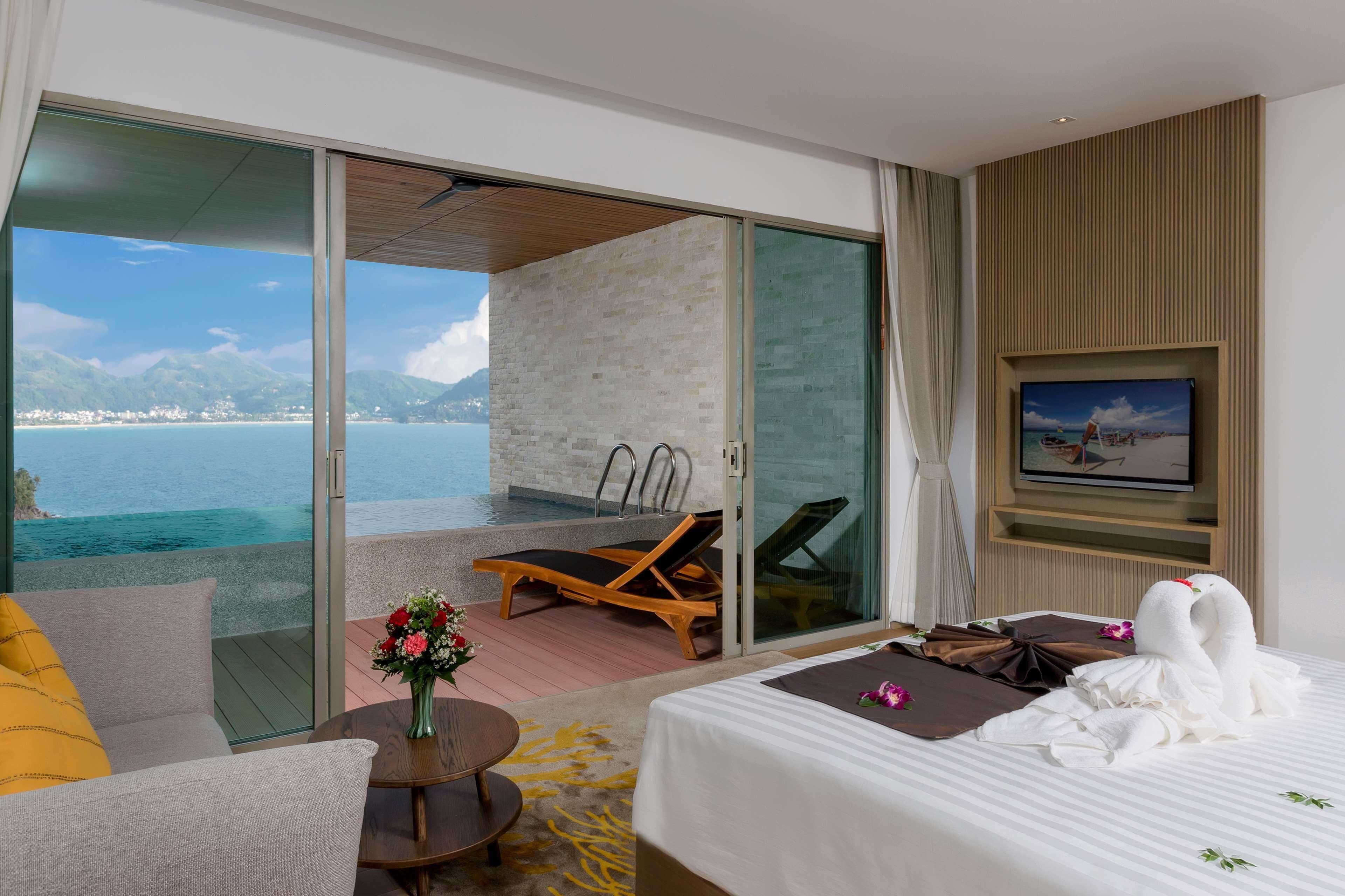 A hotel room with a bed, a sofa, a small table with a vase, a TV, and a balcony with a view of the sea and mountains.