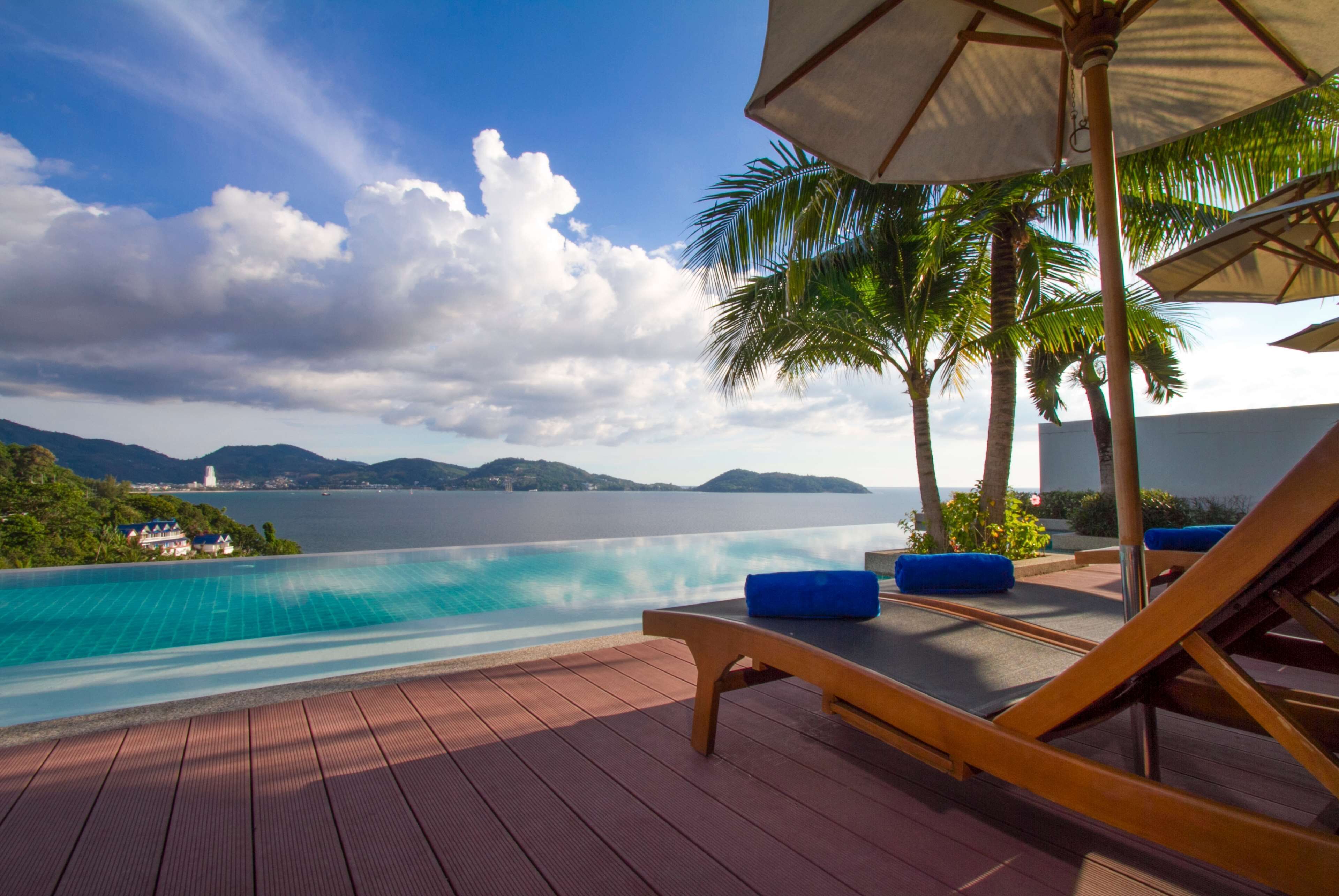 An infinity pool with a view of the sea and mountains, wooden lounge chairs, and a palm tree.
