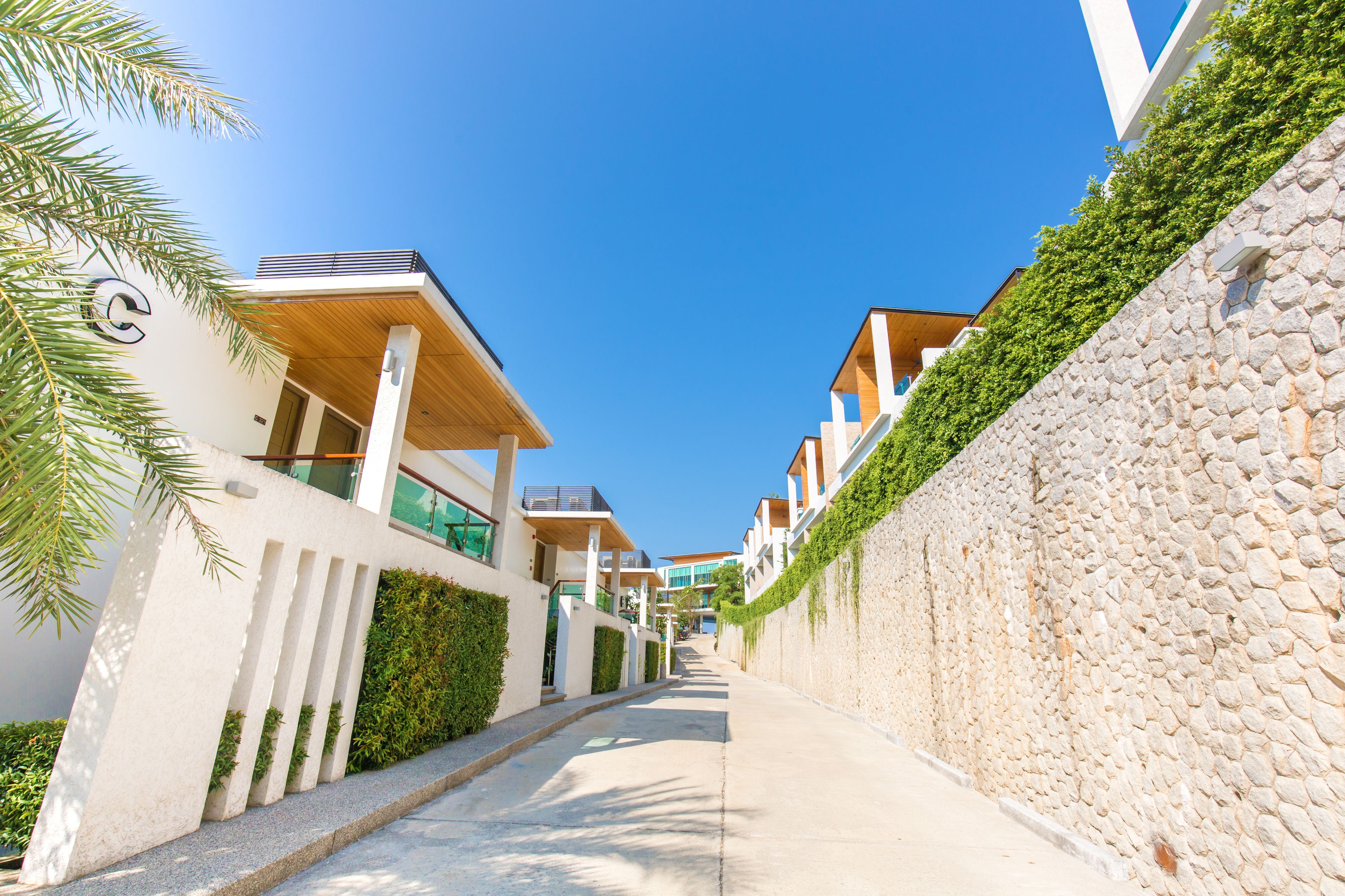 Una calle residencial moderna, con un muro de piedra y vegetación en un lado, y un edificio blanco con balcones en el otro.