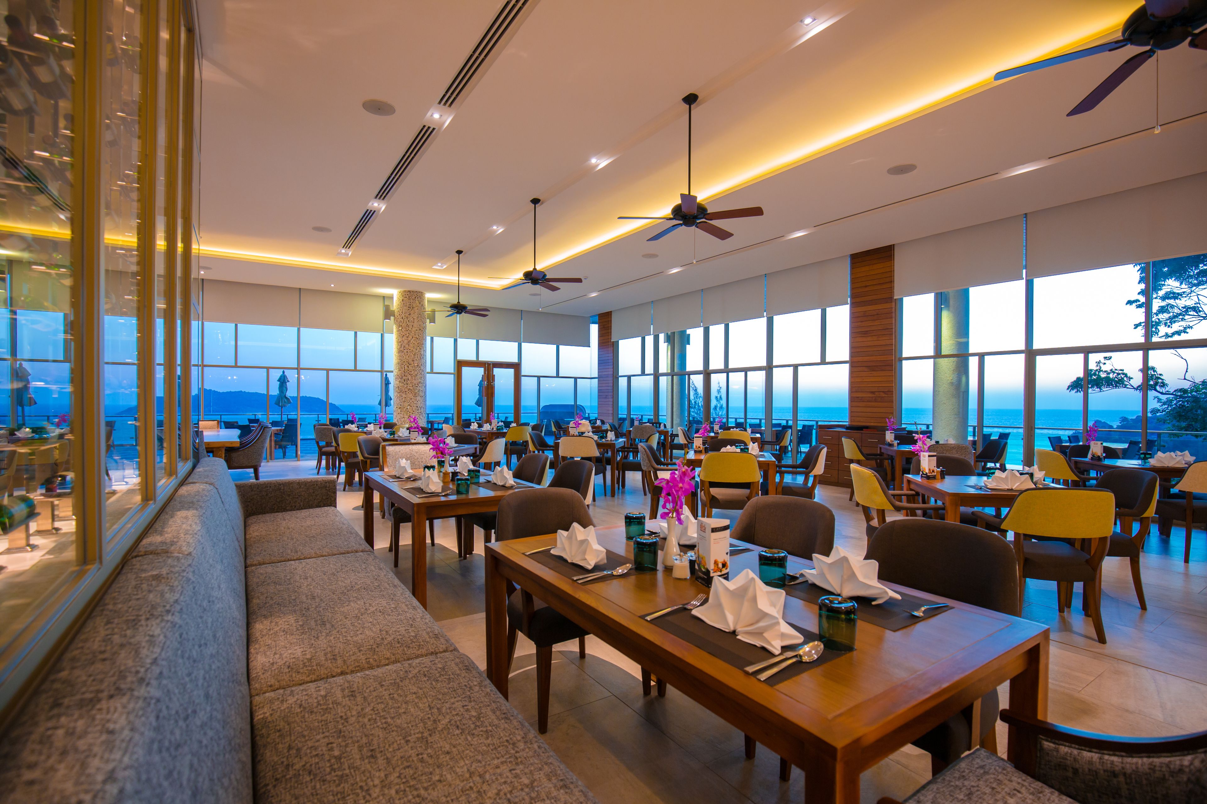 A spacious restaurant with multiple dining tables and chairs, large windows offering a view of the ocean, and ceiling fans.