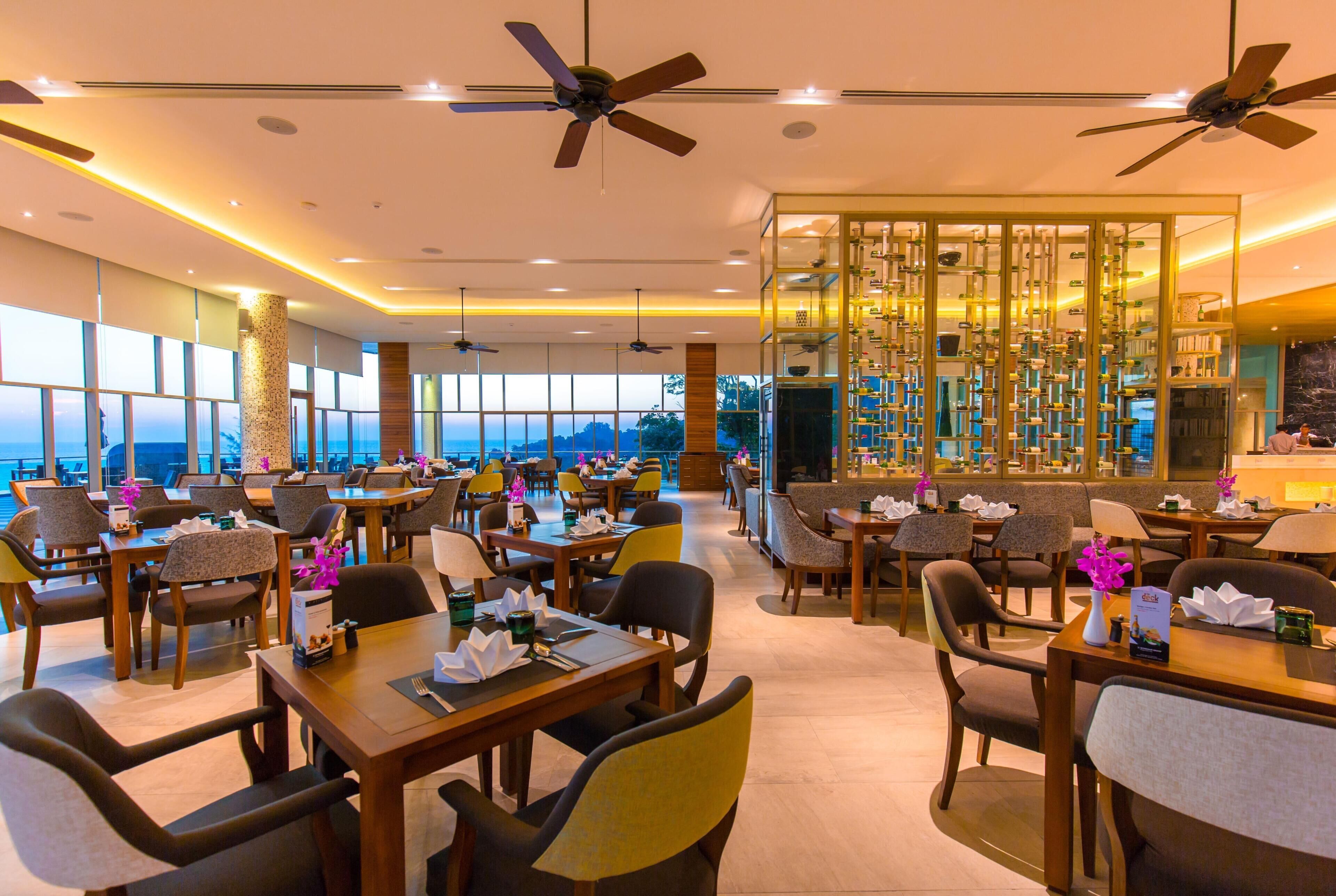A restaurant with multiple dining tables, chairs, ceiling fans, and a view of the sea.
