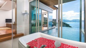 A modern bathroom with a large bathtub filled with rose petals, a glass shower enclosure, and a view of a pool and mountains.