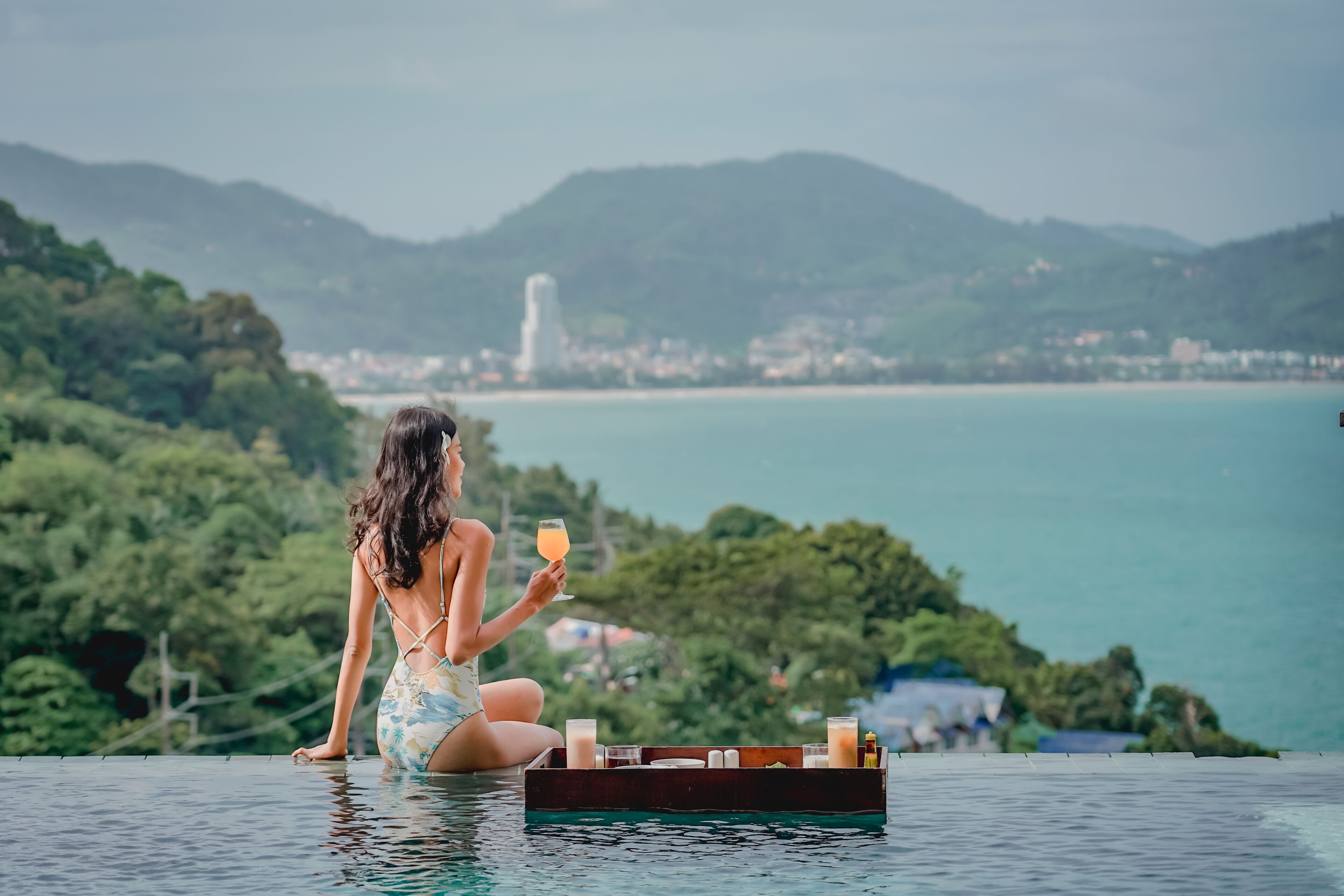 Une personne est assise au bord d’une piscine, avec un plateau rempli de boissons et de collations, et elle a devant elle un paysage côtier avec des montagnes et un phare.