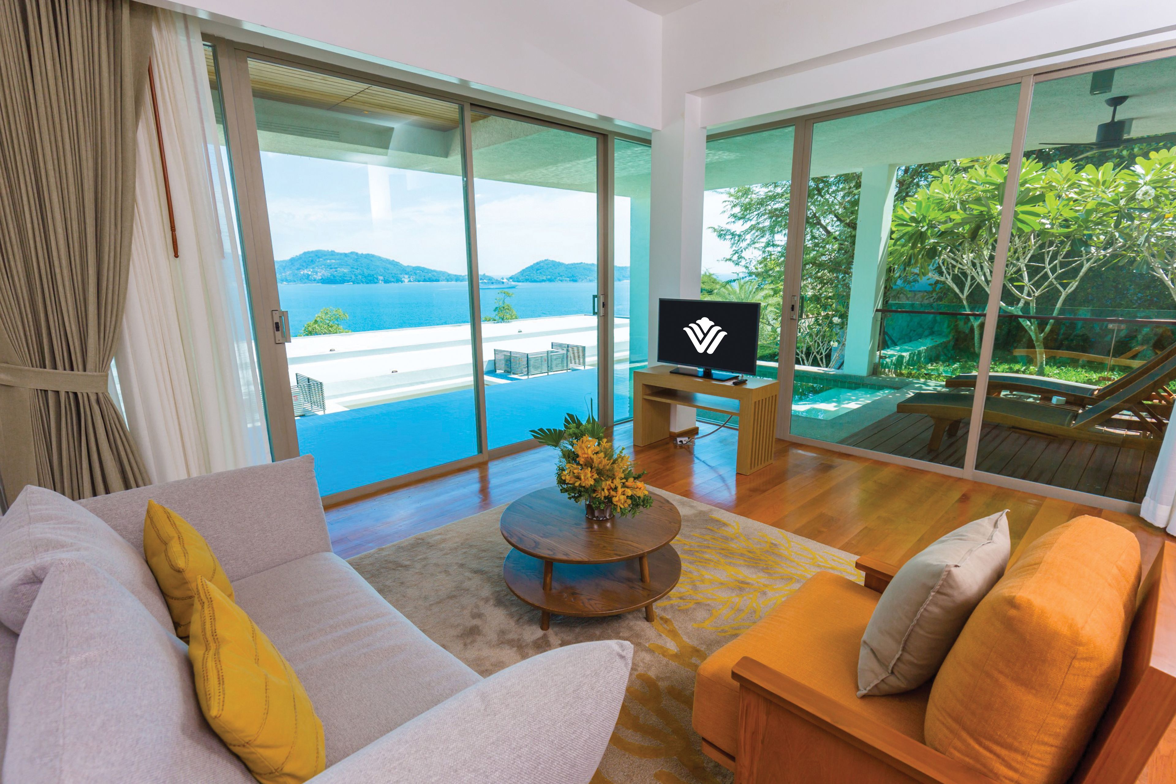 A modern living room with a sofa, a coffee table, and a TV. There is a large sliding glass door leading to a balcony with a pool and a view of the sea.