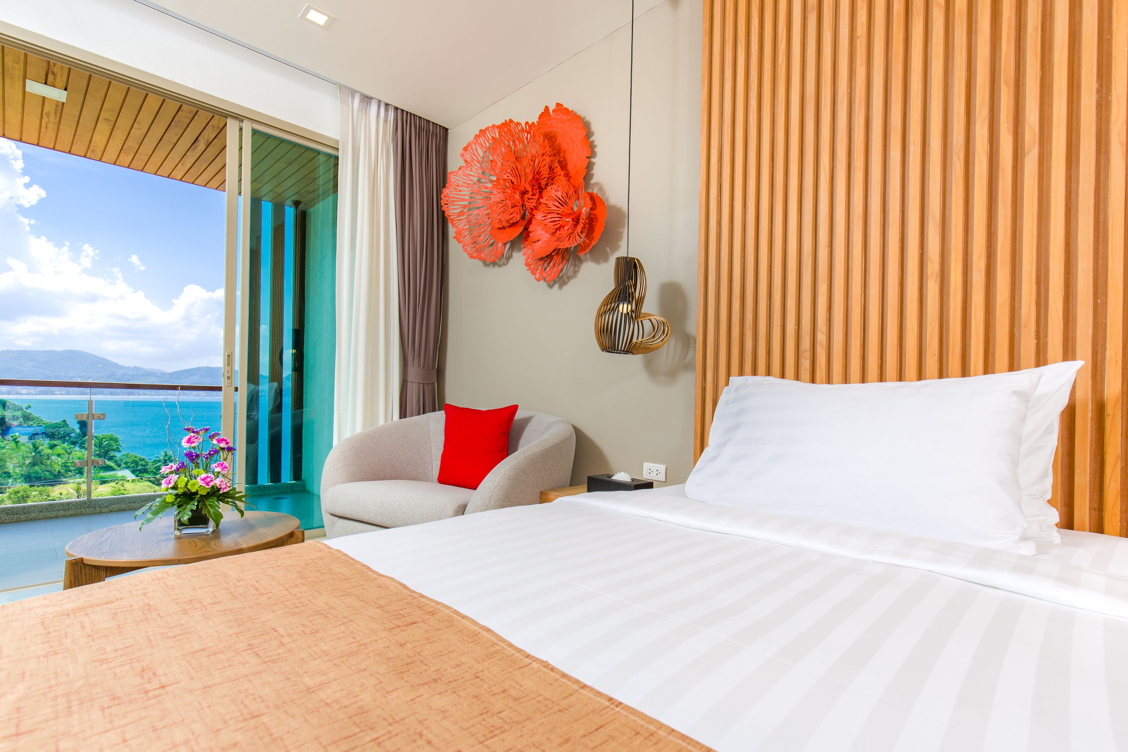 A modern hotel room with a large bed, a view of the sea, and a balcony with flowers.