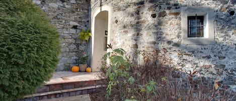 Un edificio de piedra con una ventana pequeña y una puerta, rodeado de plantas y un camino de piedra.