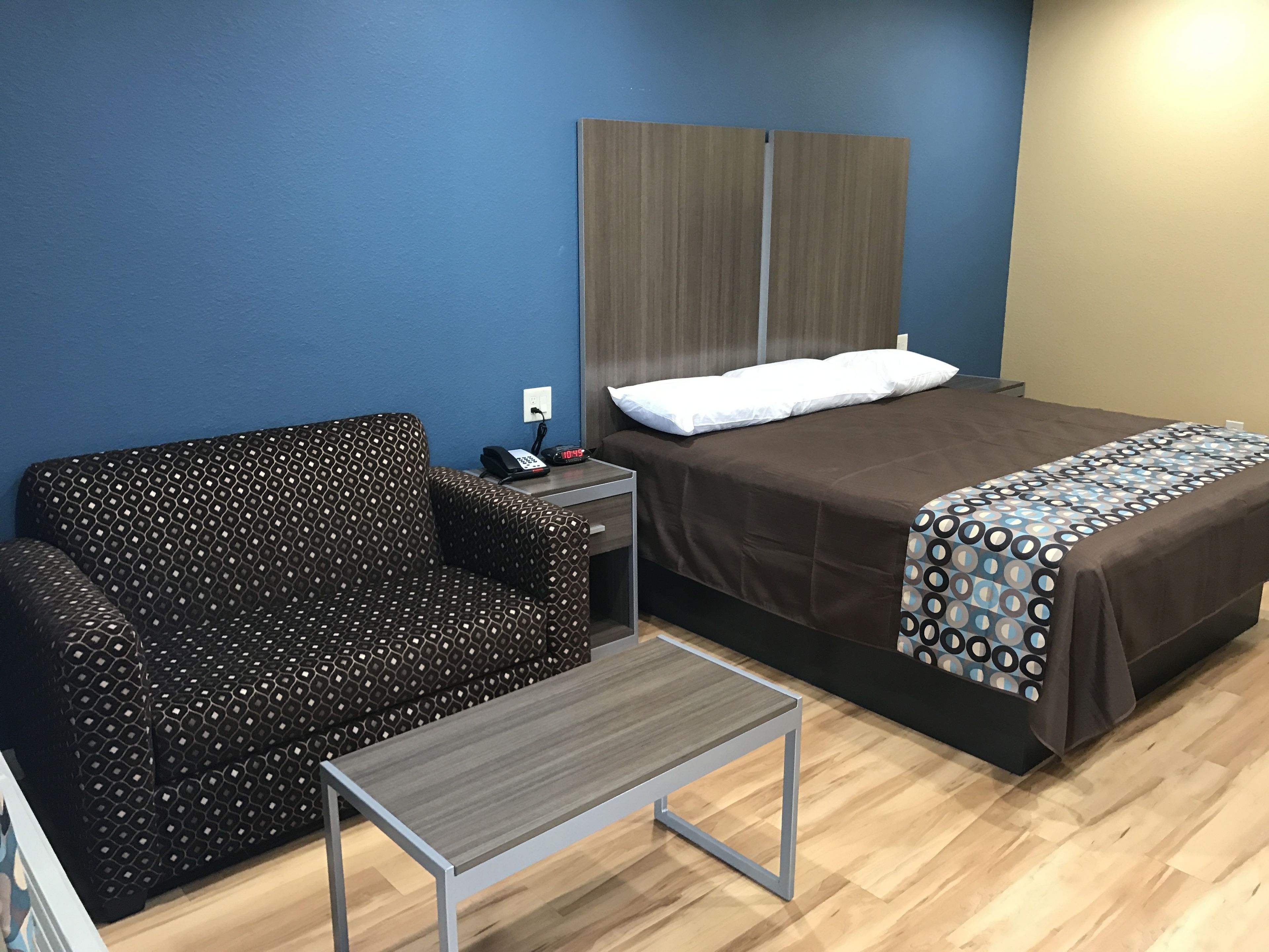 A hotel room with a bed, a sofa, a small table, and a nightstand with a phone and clock.