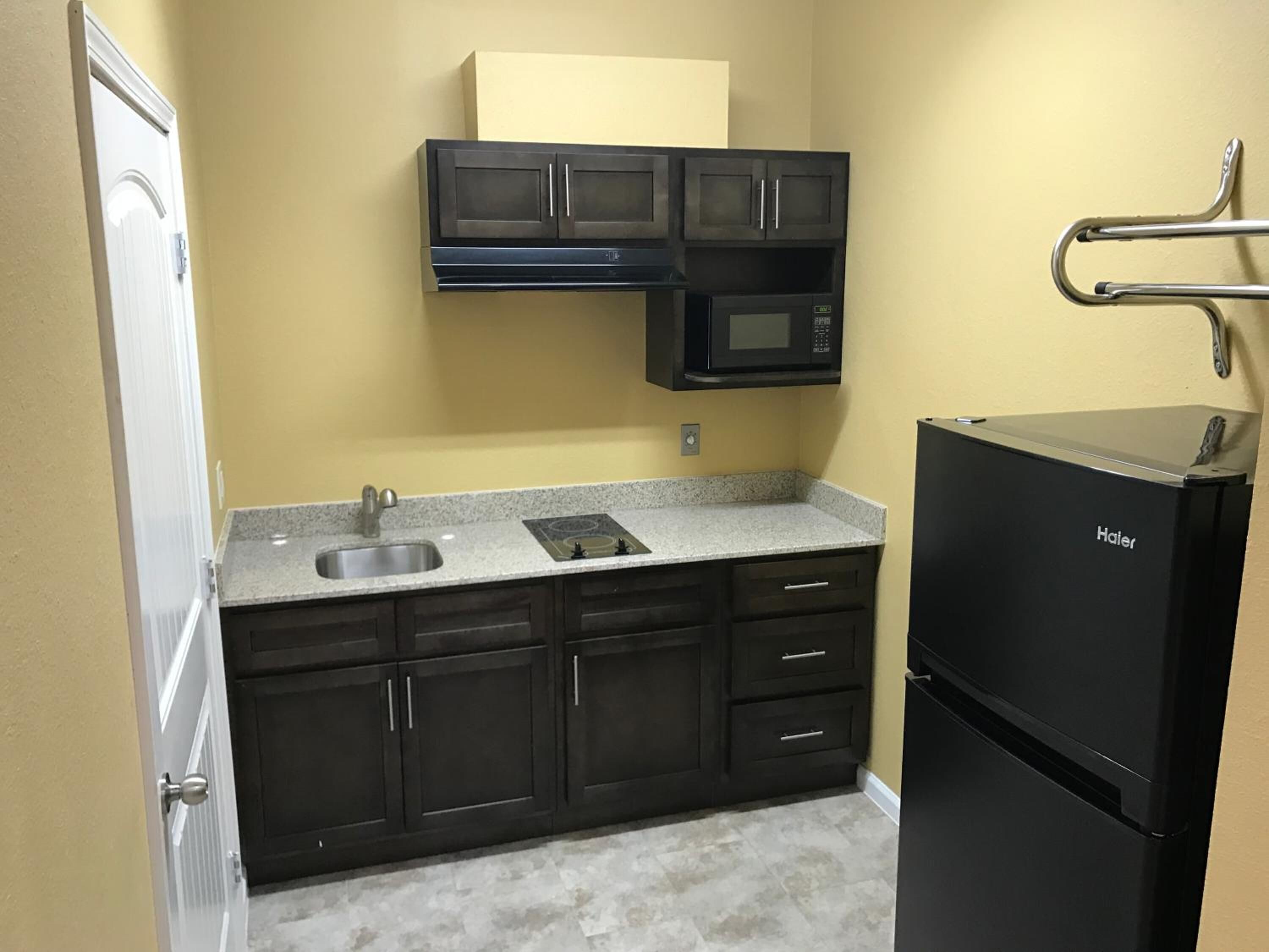 Una cocina compacta con armarios oscuros, un lavamanos de acero inoxidable, un refrigerador Haier negro y un microondas sobre la estufa.