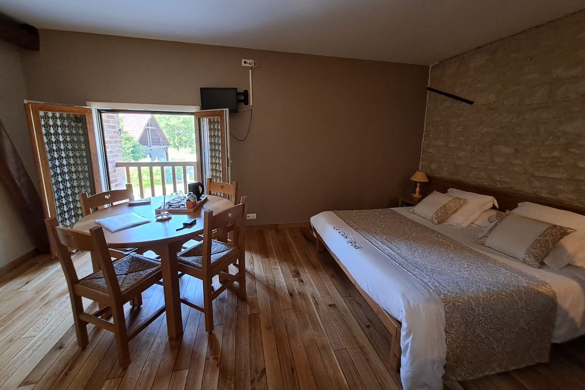 Ein Zimmer mit Holzboden, einem Bett mit gemustertem Überwurf, einem Tisch mit Stühlen und einem Fenster mit Blick auf Grünflächen.